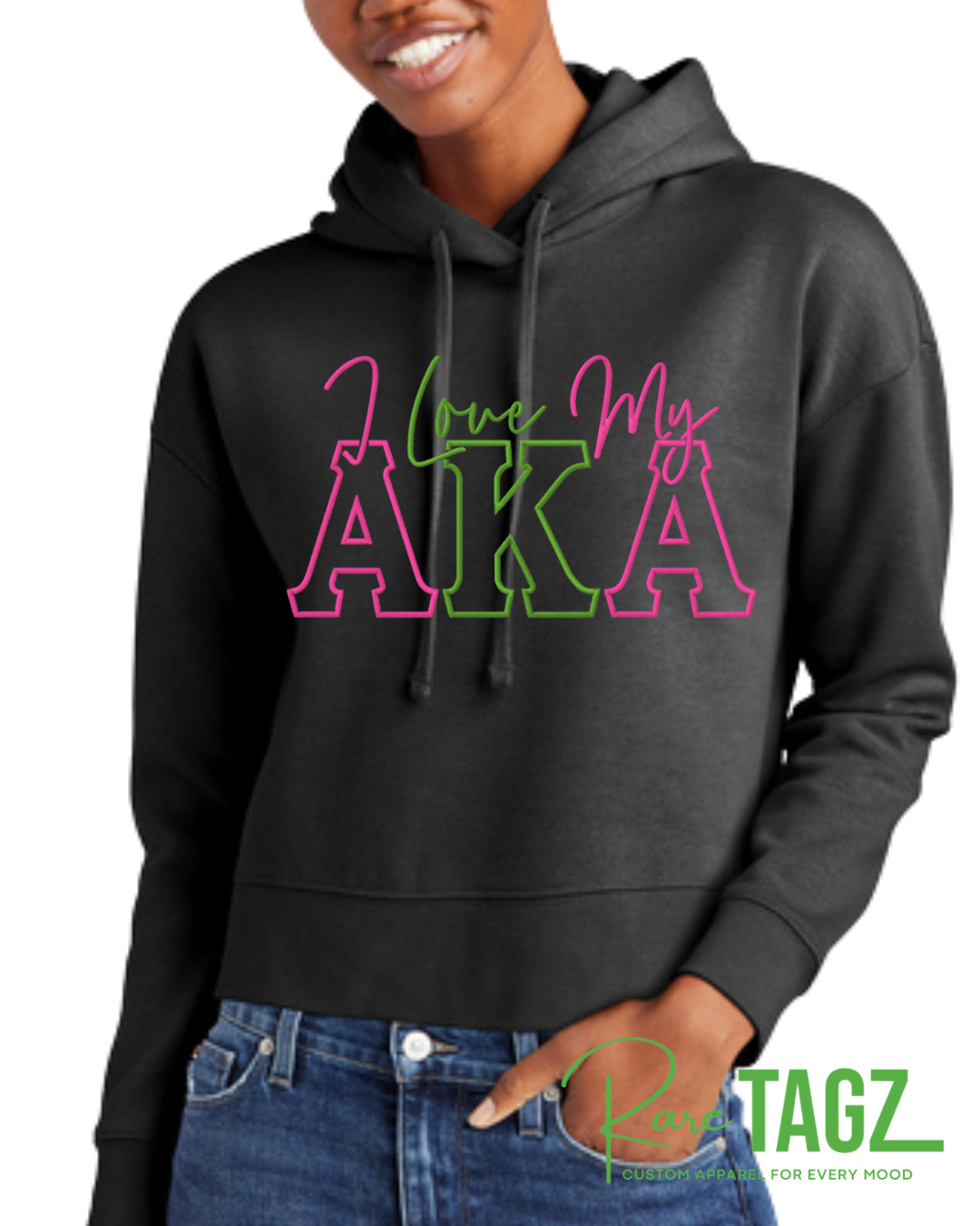 Woman wearing a black hoodie with pink and green lettering that reads ‘I Love My AKA.’ The hoodie has a relaxed fit with a kangaroo pocket and drawstring hood. Background shows colorful graffiti art. Rare Tagz logo is in the corner.