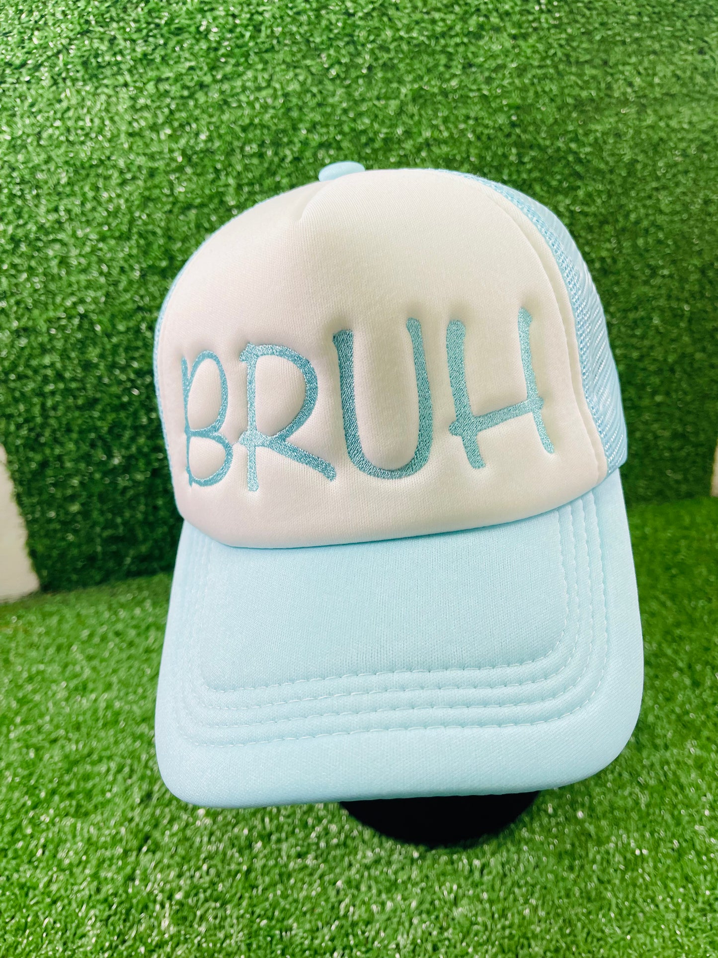 Light blue and white trucker hat with bold embroidery that reads BRUH on the front, displayed against a green turn background.