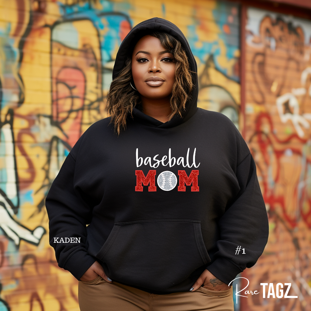 Woman wearing a custom Baseball Mom sweatshirt by Rare Tagz. A black hoodie with red and white lettering, standing against a graffiti wall. Featuring sporty outfits, bold embroidery perfect for proud baseball moms.