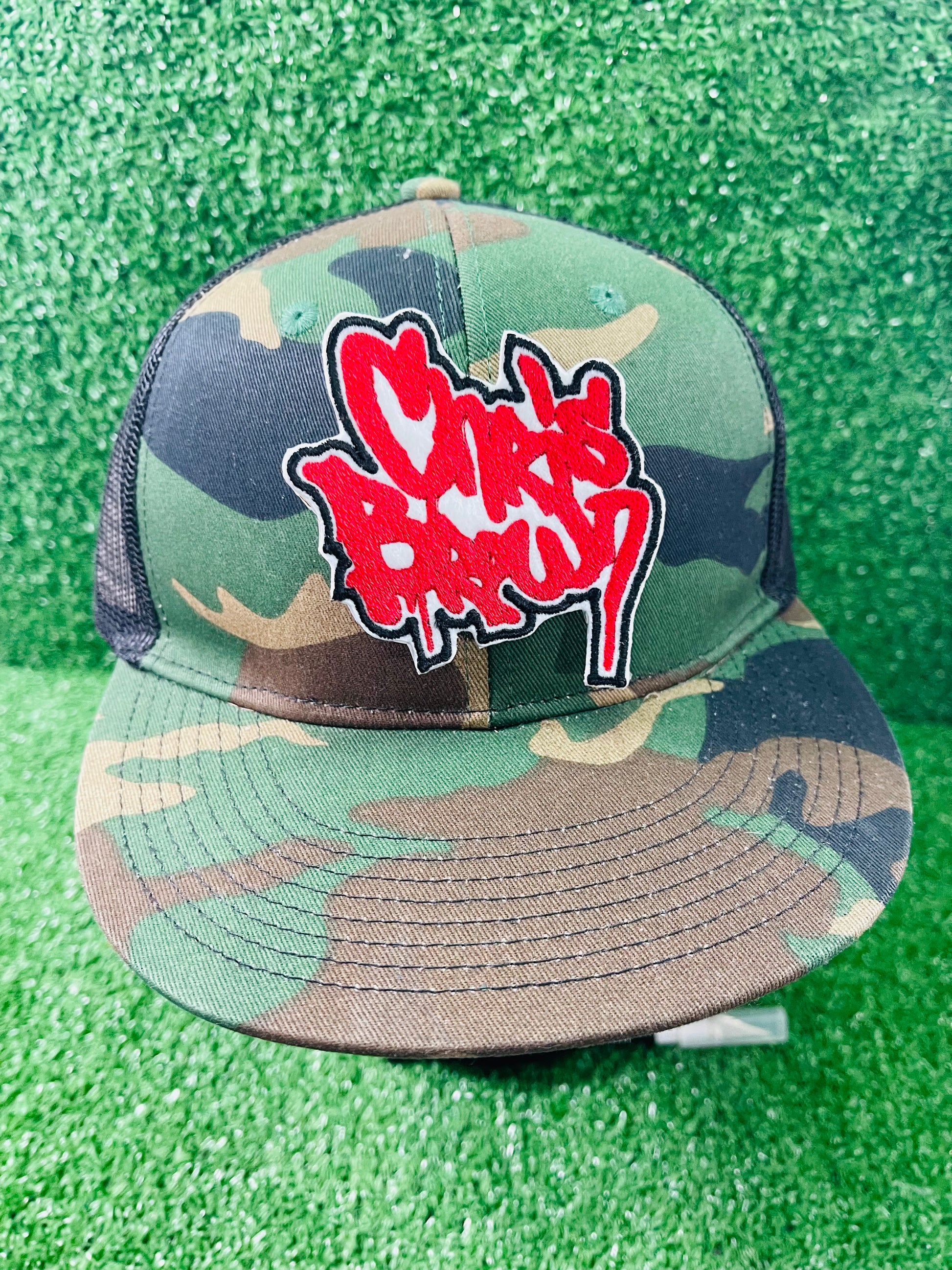 Camouflage cap with red and white logo on a green textured background. Custom-made Chris Brown inspired embroidered patches held against a vibrant green background. One patch features black graffiti-style text with a white base and gold outline; the other features red text with a white base and black outline. Designed and stitched in-house.