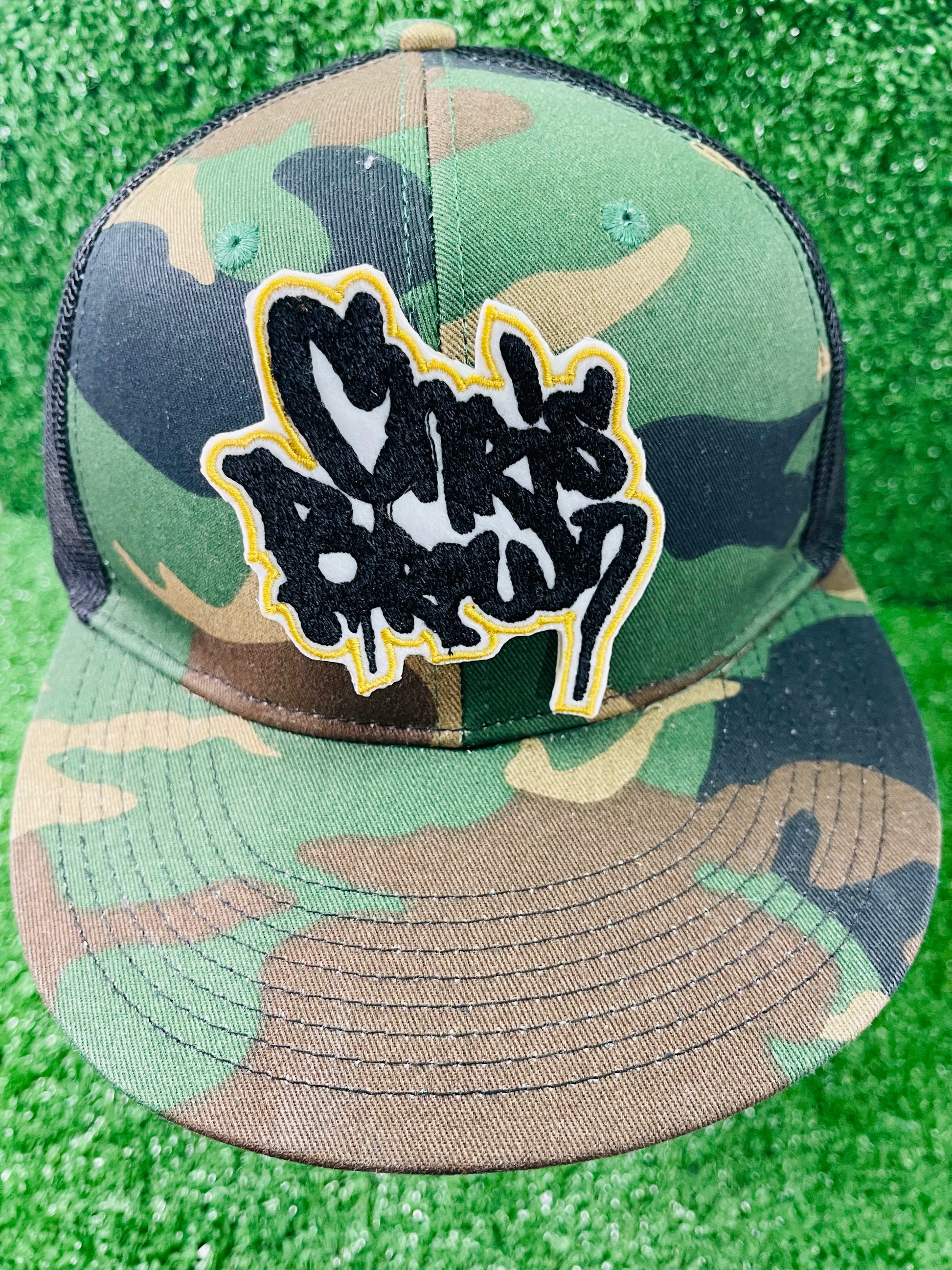 Camouflage cap with a logo on a green textured background. Two custom-made Chris Brown-inspired embroidered patches held against a vibrant green background. One patch features black graffiti-style text with a white base and gold outline; the other features red text with a white base and black outline. Designed and stitched in-house.