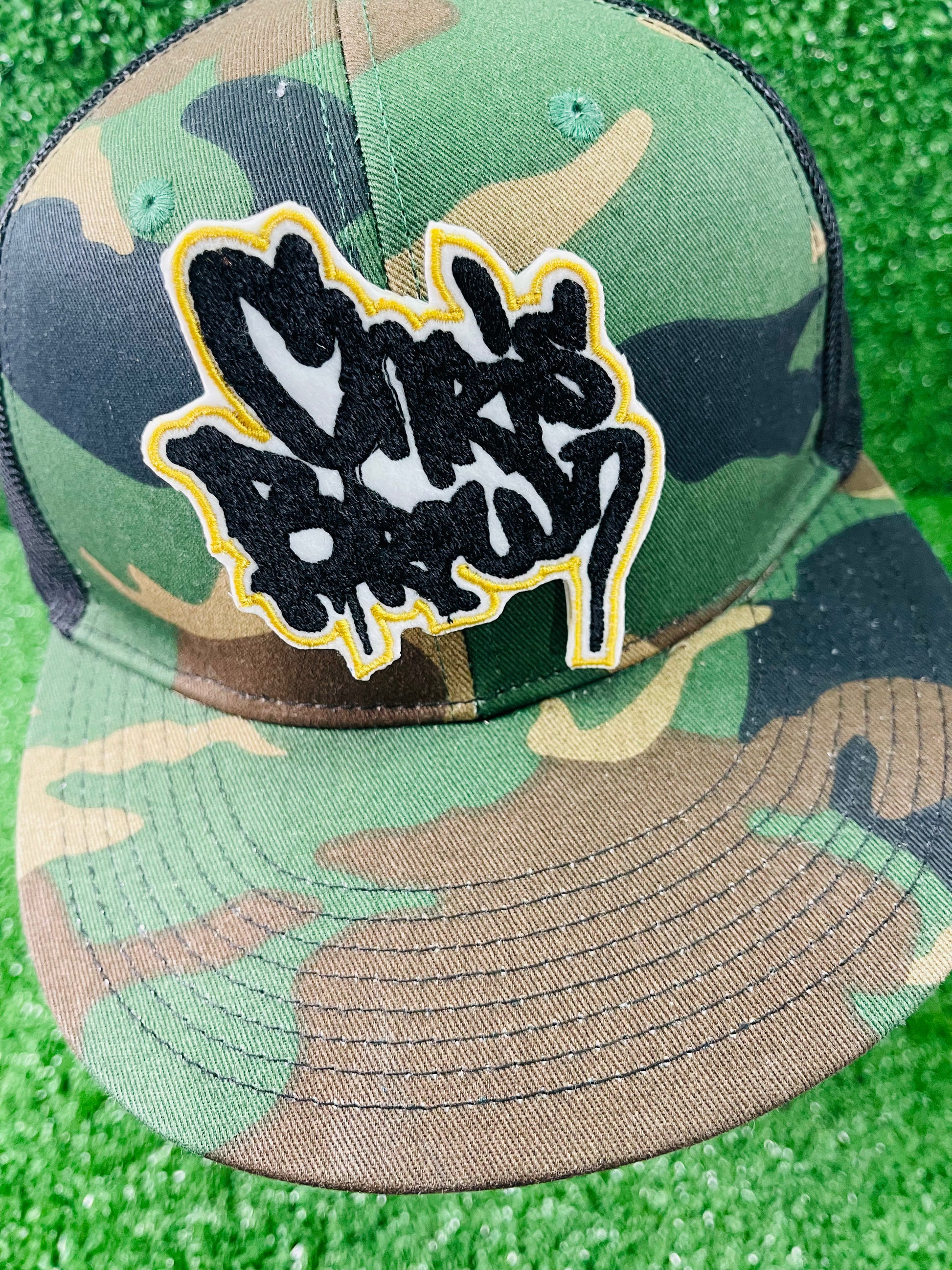 Custom-made Chris Brown-inspired embroidered patches held against a vibrant green background. One patch features black graffiti-style text with a white base and gold outline; the other features red text with a white base and black outline. Designed and stitched in-house.