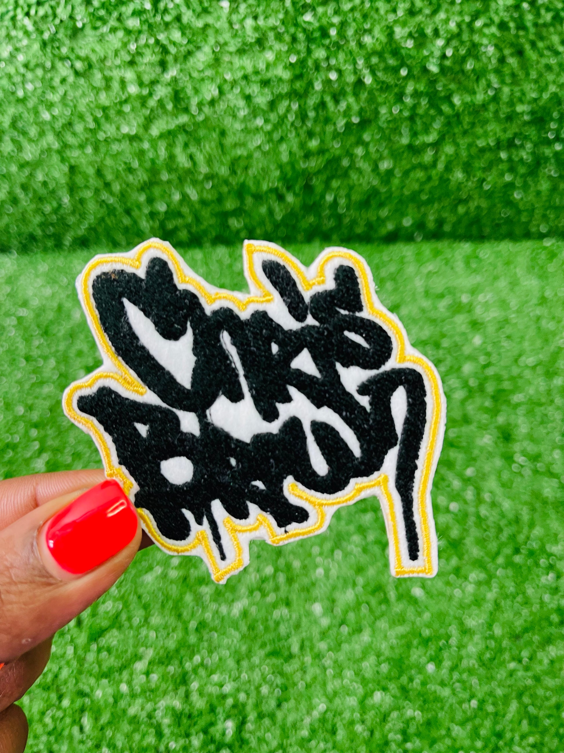 Black and white logo patch held by a hand with red nail polish against a green textured background. Custom-made Chris Brown inspired embroidered patches held against a vibrant green background. One patch features black graffiti-style text with a white base and gold outline; the other features red text with a white base and black outline. Designed and stitched in-house.