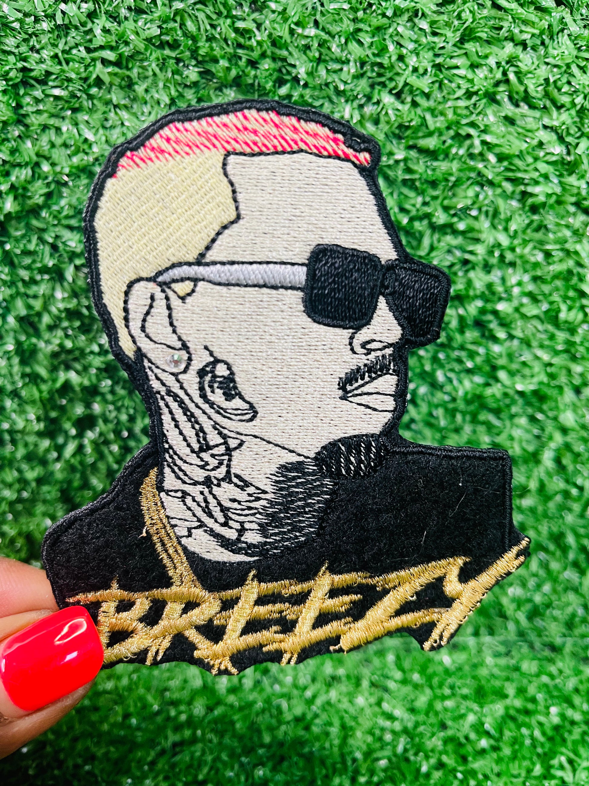 Custom embroidered patch featuring chris brown's side profile with blonde and pink hair, black sunglasses and detailed tattoos, with the word Breezy stitched in gold thread beneath the design, Camouflage cap with a graphic design and 'Breezy' text on a green surface