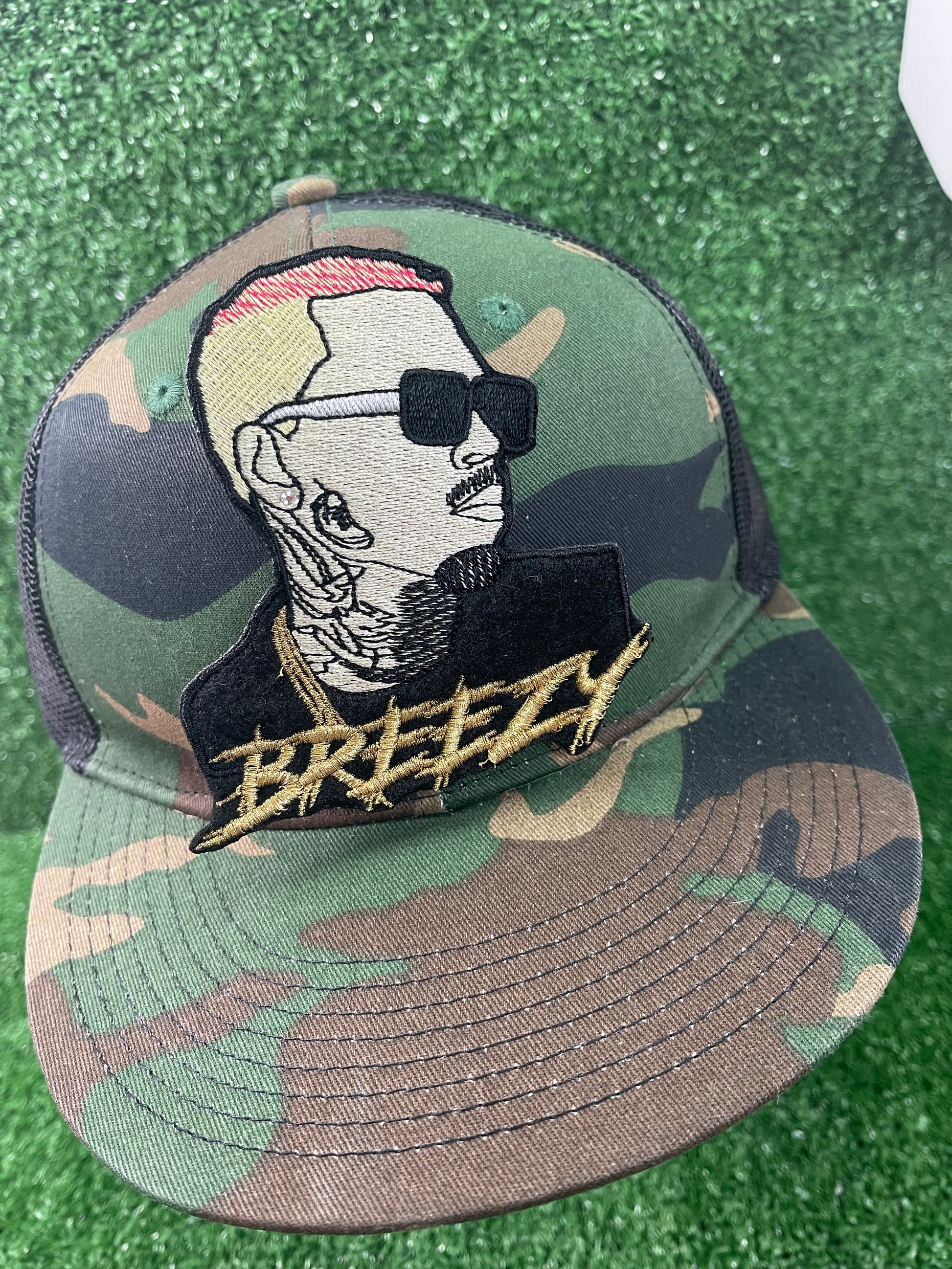 Custom embroidered patch featuring chris brown's side profile with blonde and pink hair, black sunglasses and detailed tattoos, with the word Breezy stitched in gold thread beneath the design, Camouflage cap with a graphic design and 'Breezy' text on a green surface
