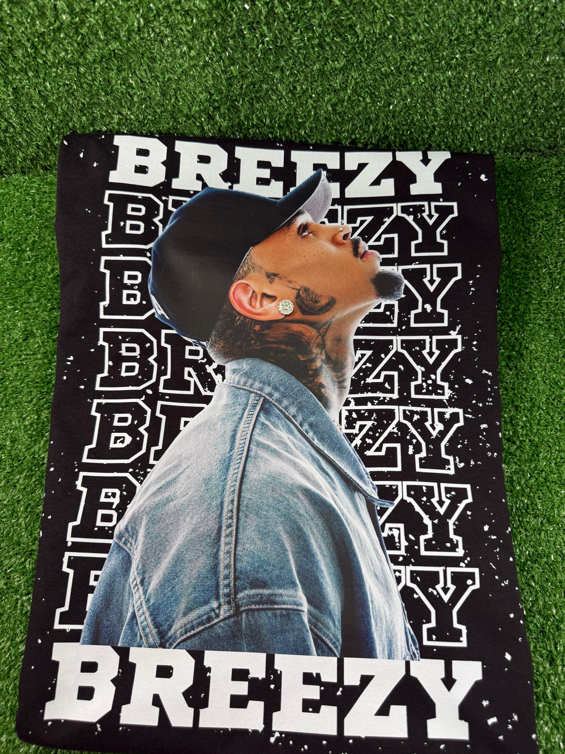 Chris Brown wearing a denim jacket with 'BREEZY' text on a green grass background. Black Breezy Bowl tour T-shirt with bold white lettering and Chris Brown graphic design.