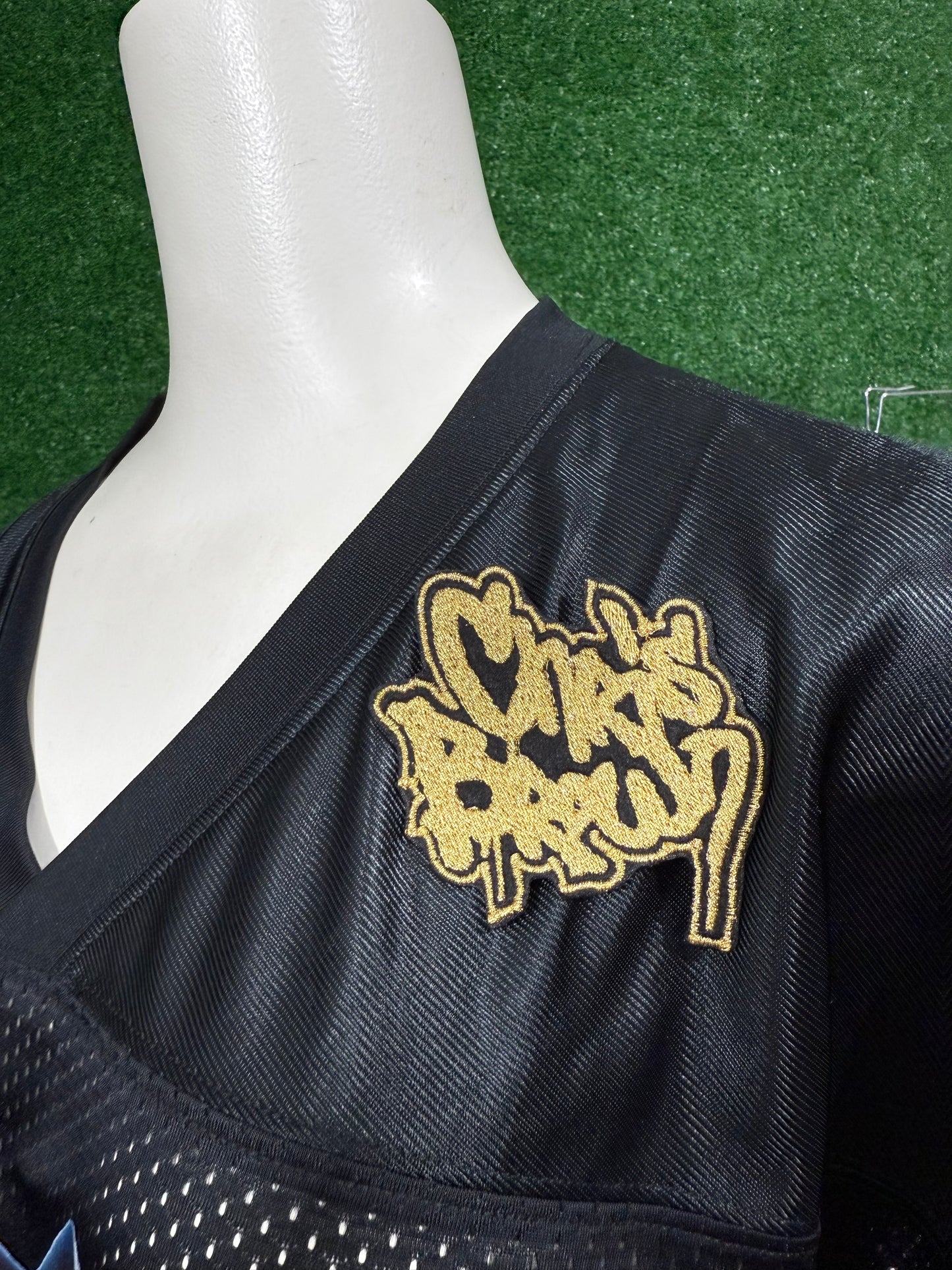Black sports jersey with gold logo on a mannequin against a green background. Custom-made Chris Brown-inspired embroidered patches held against a vibrant green background. One patch features black graffiti-style text with a white base and gold outline; the other features red text with a white base and black outline. Designed and stitched in-house.