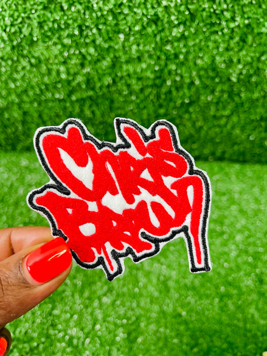 Two custom-made Chris Brown inspired embroidered patches held against a vibrant green background. One patch features black graffiti-style text with a white base and gold outline; the other features red text with a white base and black outline. Designed and stitched in-house.