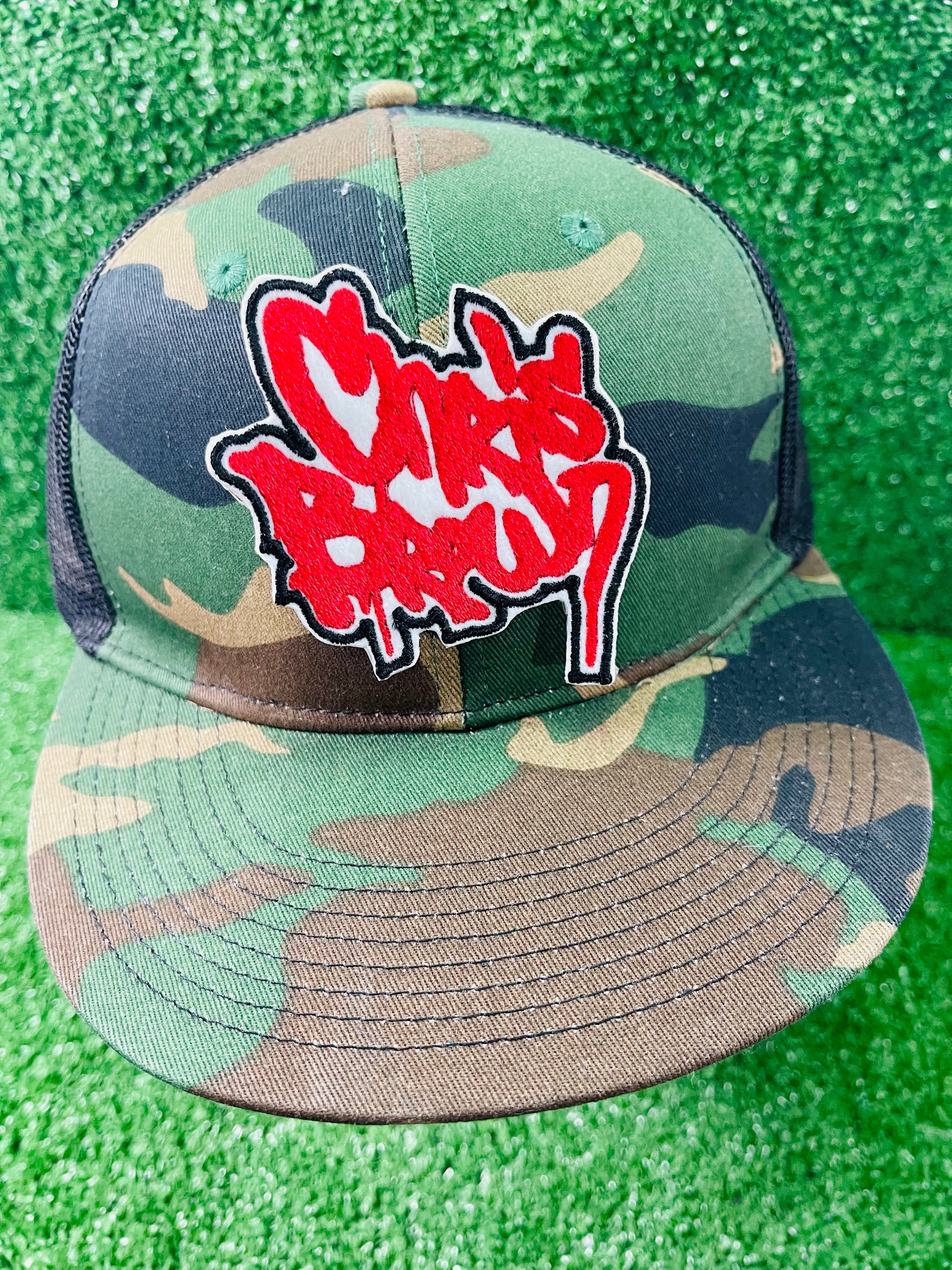 Camouflage baseball cap with red and white logo on a green grass background. custom-made Chris Brown-inspired embroidered patches held against a vibrant green background. One patch features black graffiti-style text with a white base and gold outline; the other features red text with a white base and black outline. Designed and stitched in-house.