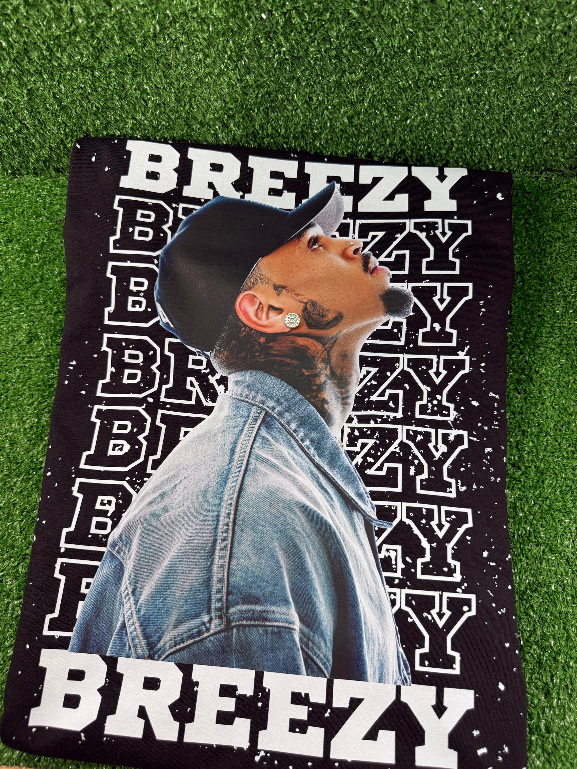 Chris Brown wearing a denim jacket with 'BREEZY' text on a green grass background. Black Breezy Bowl tour T-shirt with bold white lettering and Chris Brown graphic design.