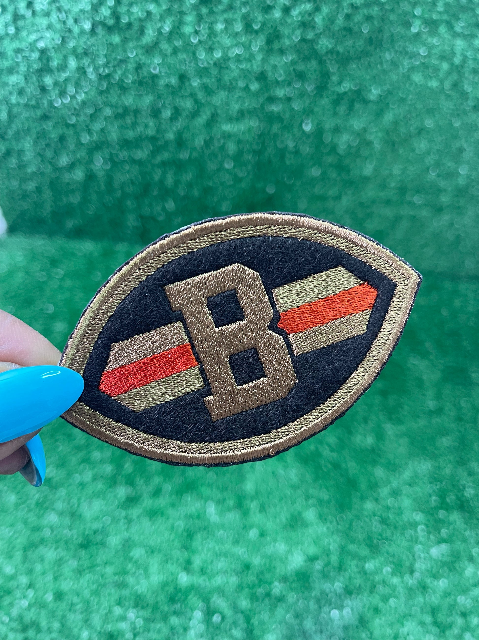 Close-up of embroidered patch shaped like a football with a Brown B in the center, dark brown background, and orange/gold stripes across each side.