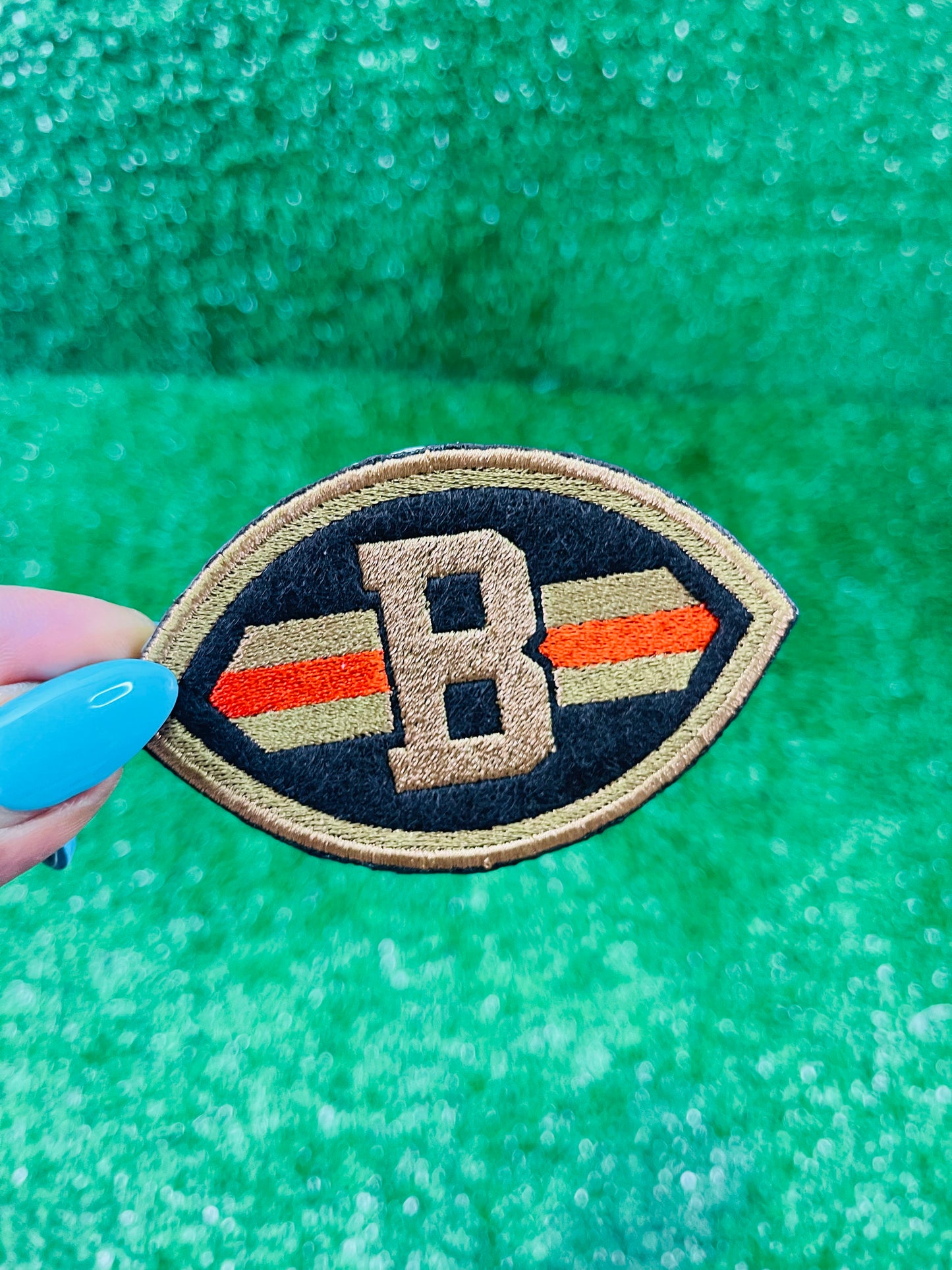 Close-up of embroidered patch shaped like a football with a Brown B in the center, dark brown background, and orange/gold stripes across each side.