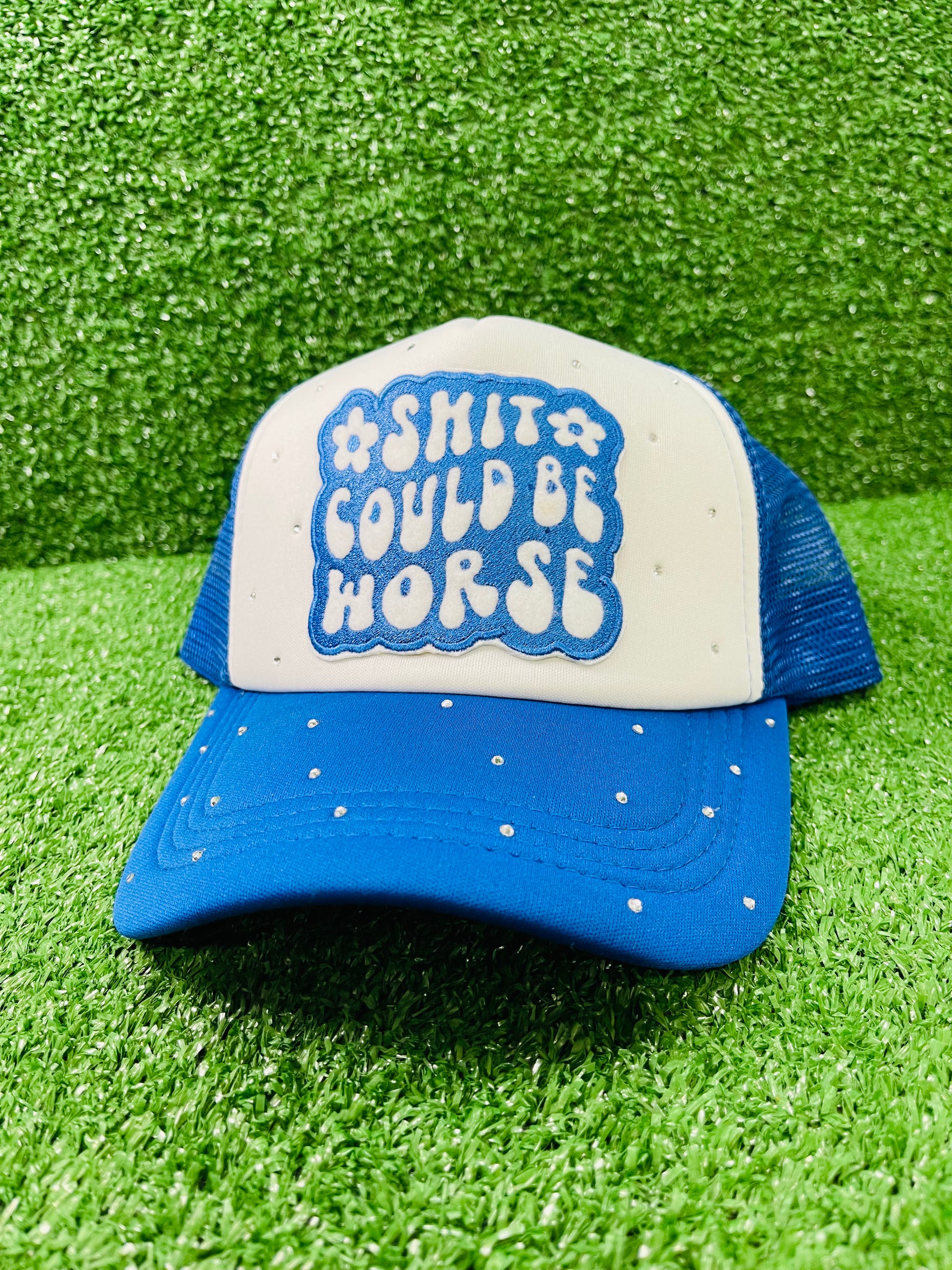 Blue and White custom trucker hat with a bold embroidered patch that reads Shit Could Be Worse and hand-placed rhinestone embellishments on the crown and brim, displayed on a green turf background.