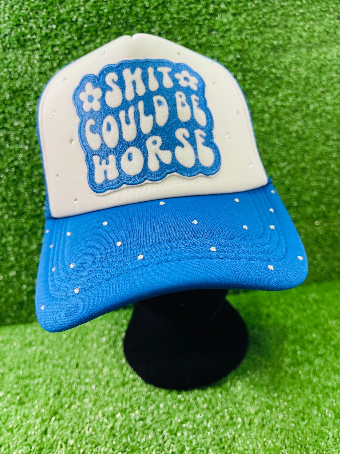Blue and White custom trucker hat with a bold embroidered patch that reads Shit Could Be Worse and hand-placed rhinestone embellishments on the crown and brim, displayed on a green turf background.