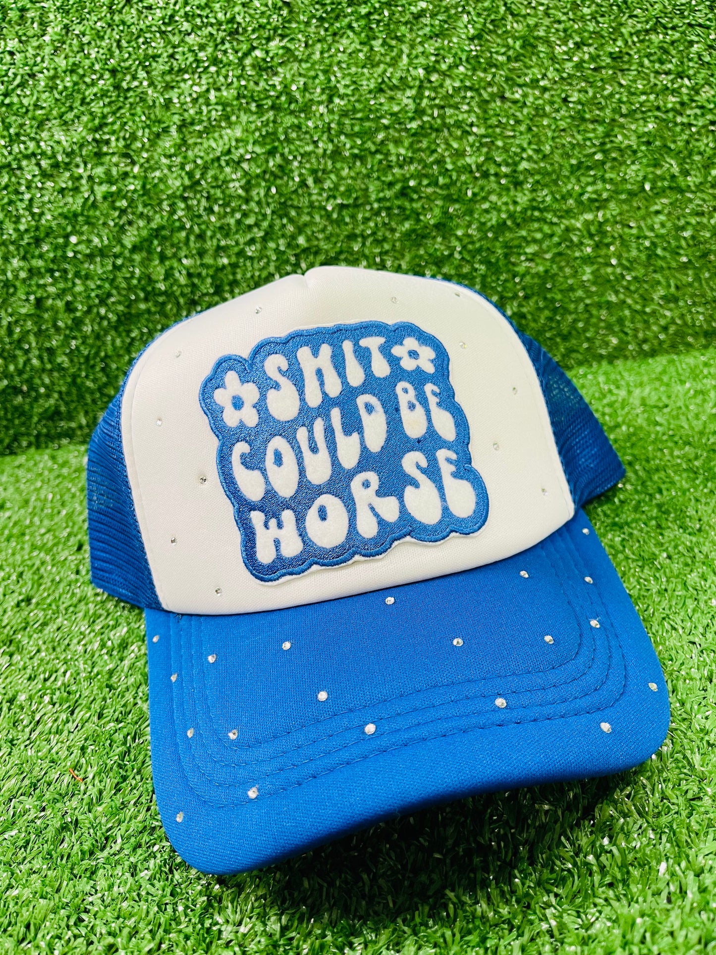Blue and White custom trucker hat with a bold embroidered patch that reads Shit Could Be Worse and hand-placed rhinestone embellishments on the crown and brim, displayed on a green turf background.