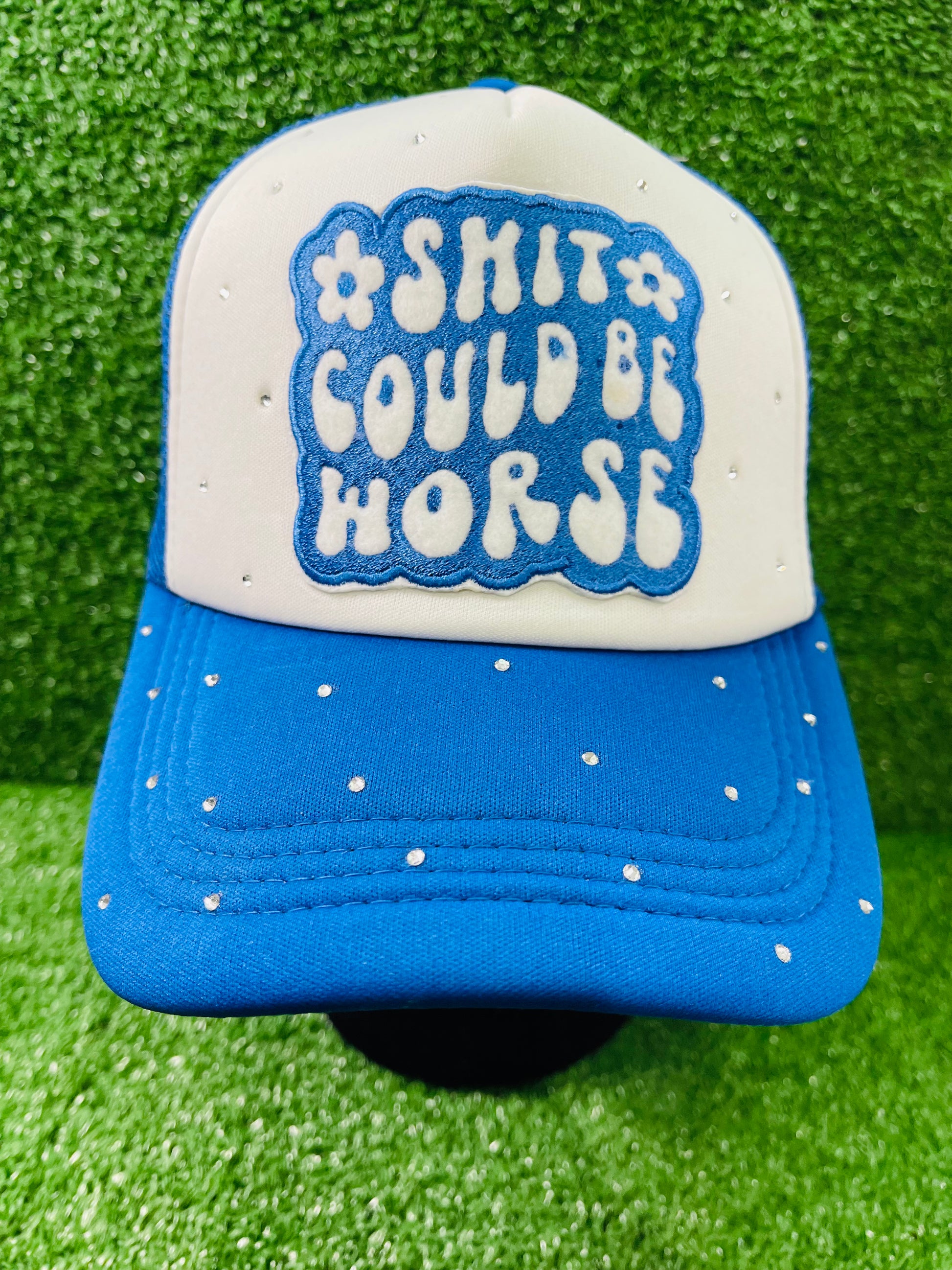 Blue and White custom trucker hat with a bold embroidered patch that reads Shit Could Be Worse and hand-placed rhinestone embellishments on the crown and brim, displayed on a green turf background.