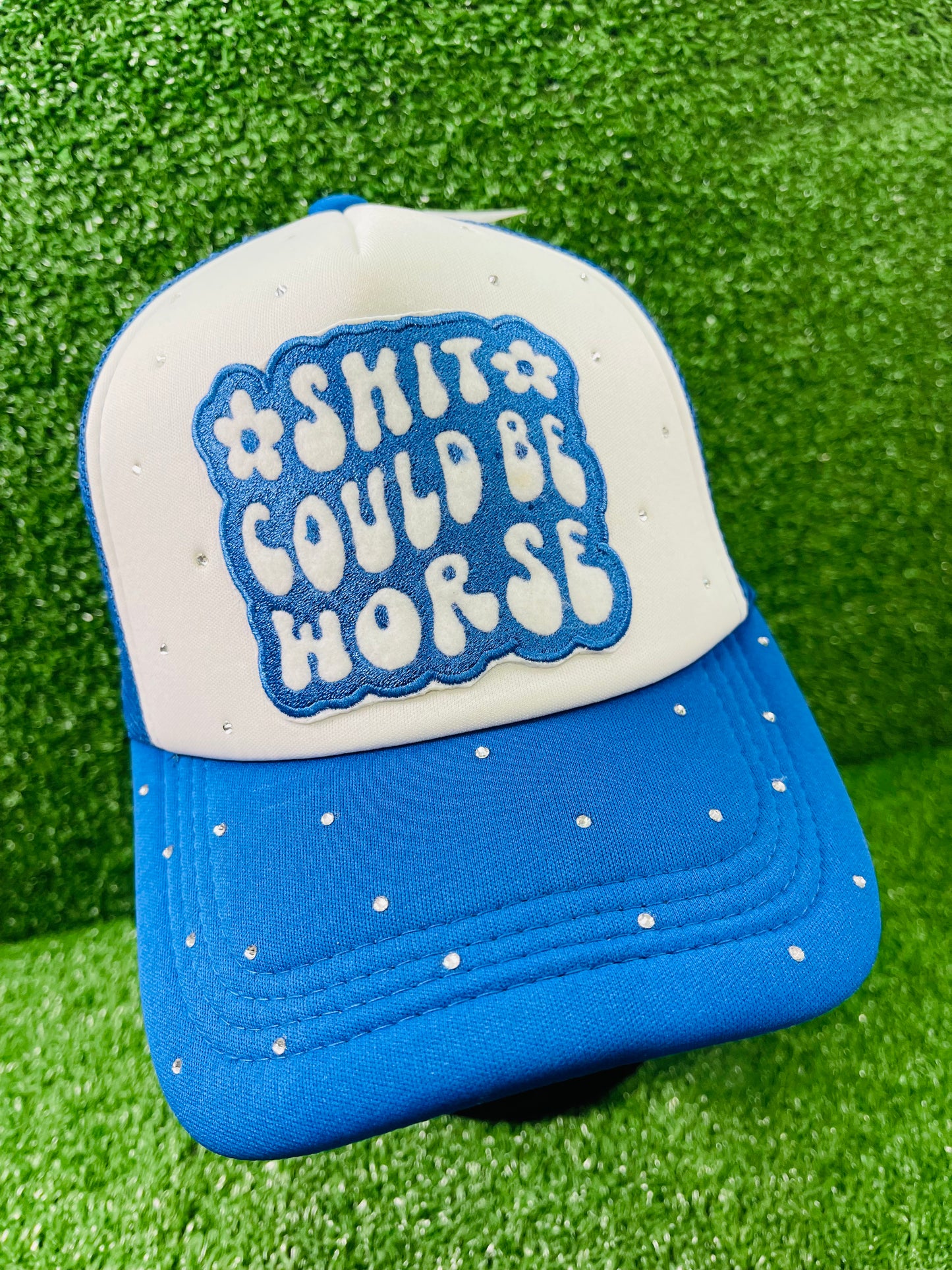 Blue and White custom trucker hat with a bold embroidered patch that reads Shit Could Be Worse and hand-placed rhinestone embellishments on the crown and brim, displayed on a green turf background.