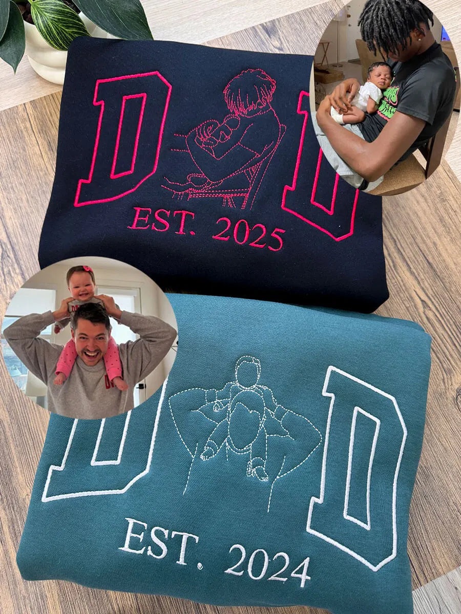 Two folded sweatshirts with embroidered custom designs of fathers with their babies. One black sweatshirt reads DAD EST 2025 with red stitching od a dad holding a newborn. The second, in teal, says DAD EST 2024 with a white thread showing a dad giving a child a piggy back ride.