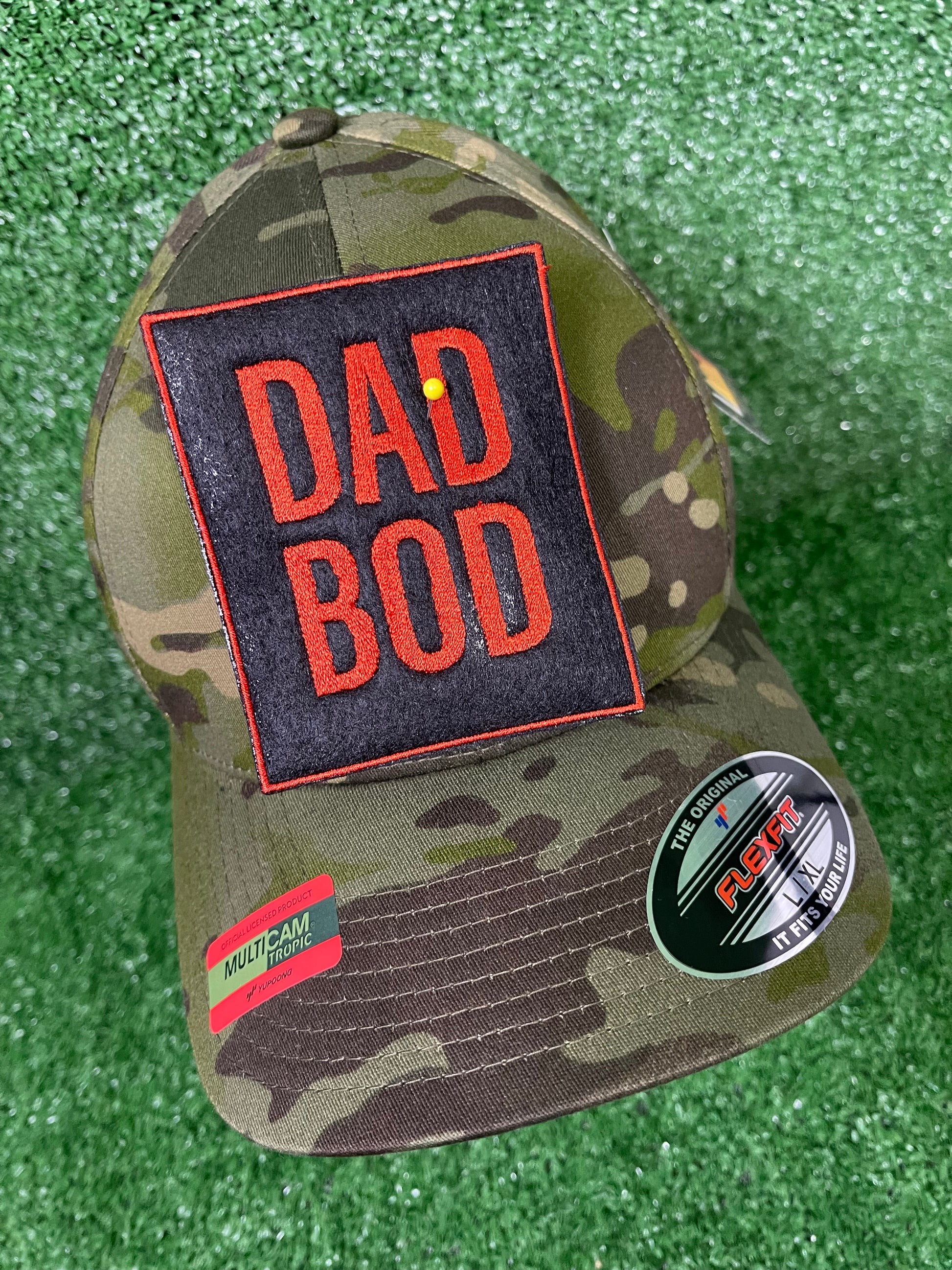 Camo trucker hat with large embroidered Dad Bod patch in bold burnt orange letters on a black background, displayed on green turf. Patch is iron-on and embroidered for Father's Day or Dad themed apparel.