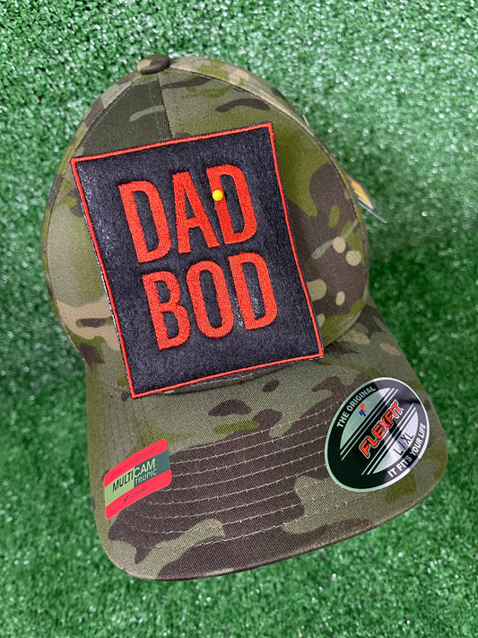 Camo trucker hat with large embroidered Dad Bod patch in bold burnt orange letters on a black background, displayed on green turf. Patch is iron-on and embroidered for Father's Day or Dad themed apparel.