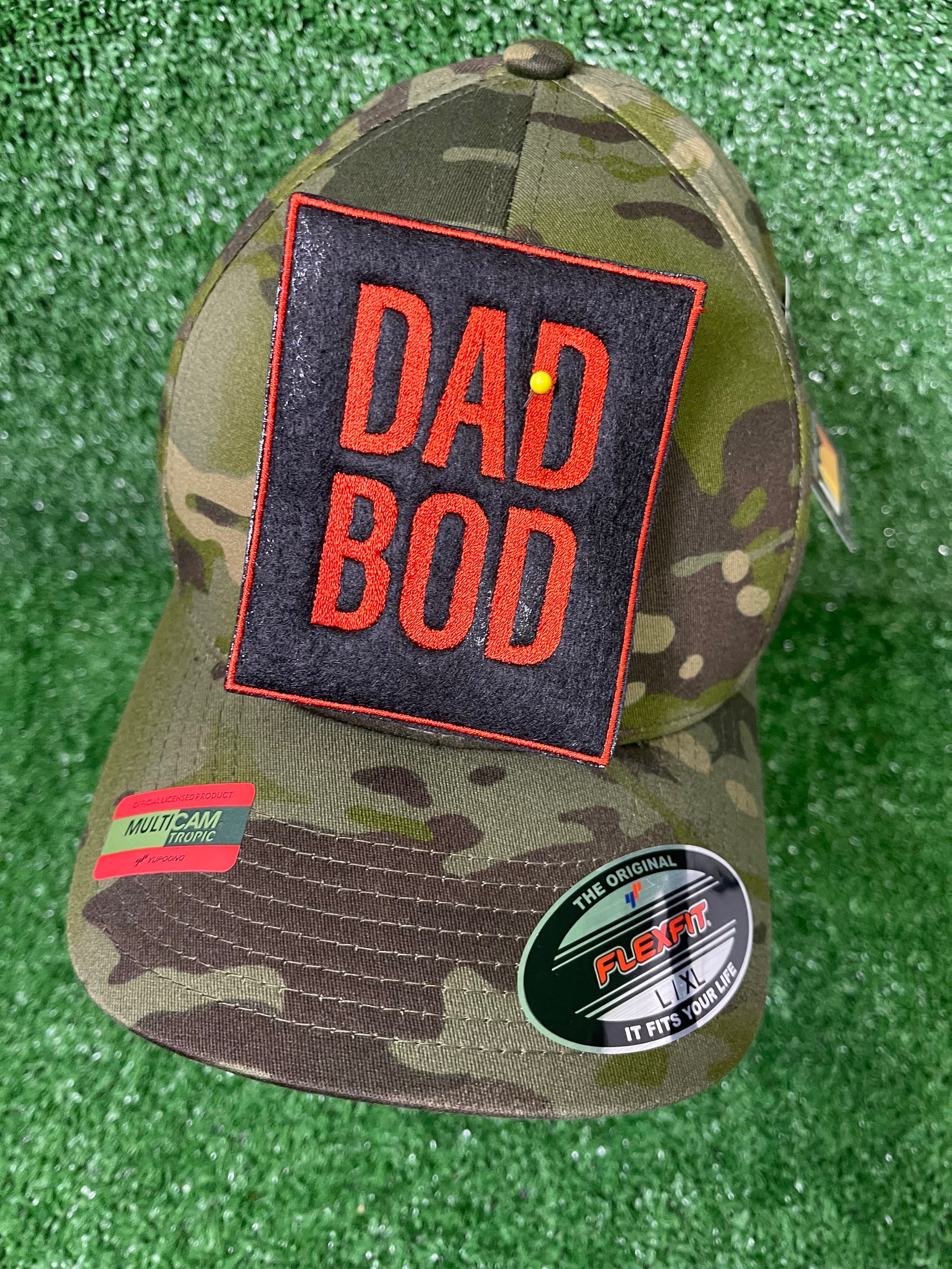 Camo trucker hat with large embroidered Dad Bod patch in bold burnt orange letters on a black background, displayed on green turf. Patch is iron-on and embroidered for Father's Day or Dad themed apparel.