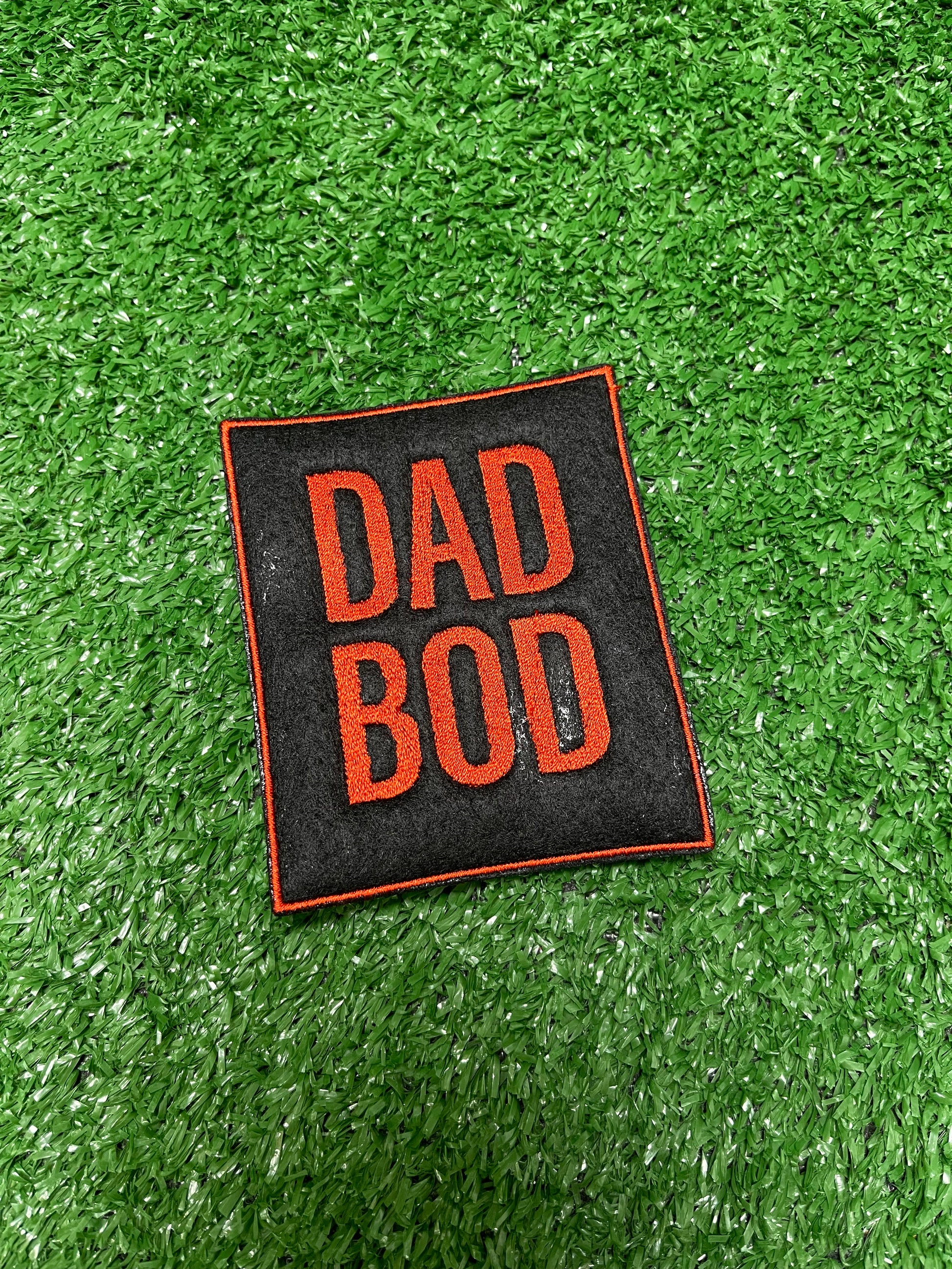 Large embroidered Dad Bod patch in bold burnt orange letters on a black background, displayed on green turf. Patch is iron-on and embroidered for Father's Day or Dad themed apparel.