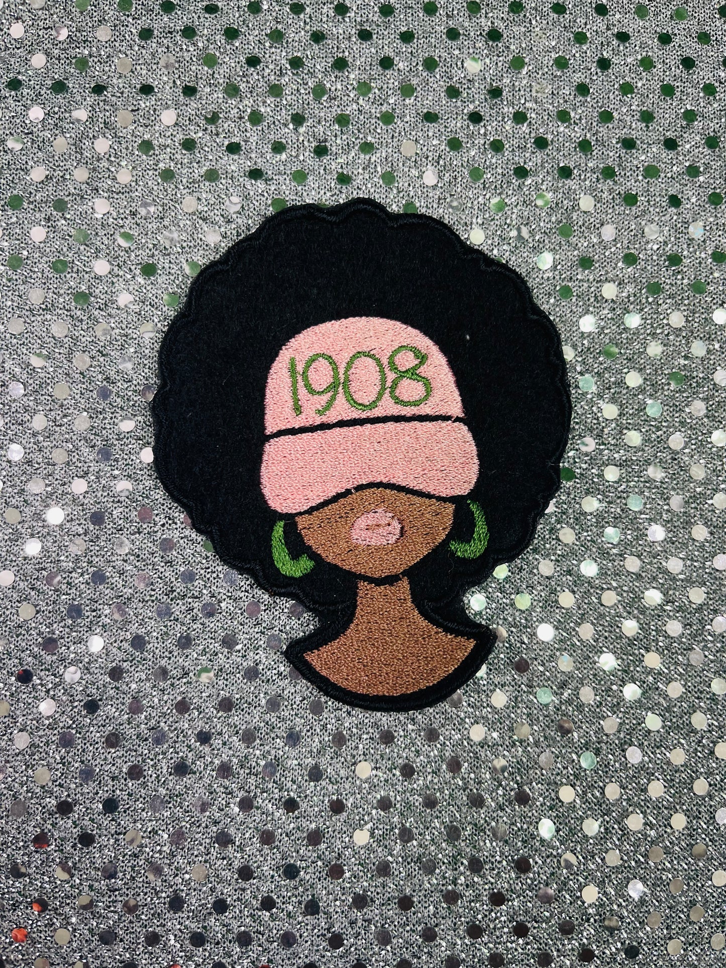 Patch of a woman with an afro, wearing a pink headscarf with '1908' text, on a textured gray background. Alpha Kappa Alpha