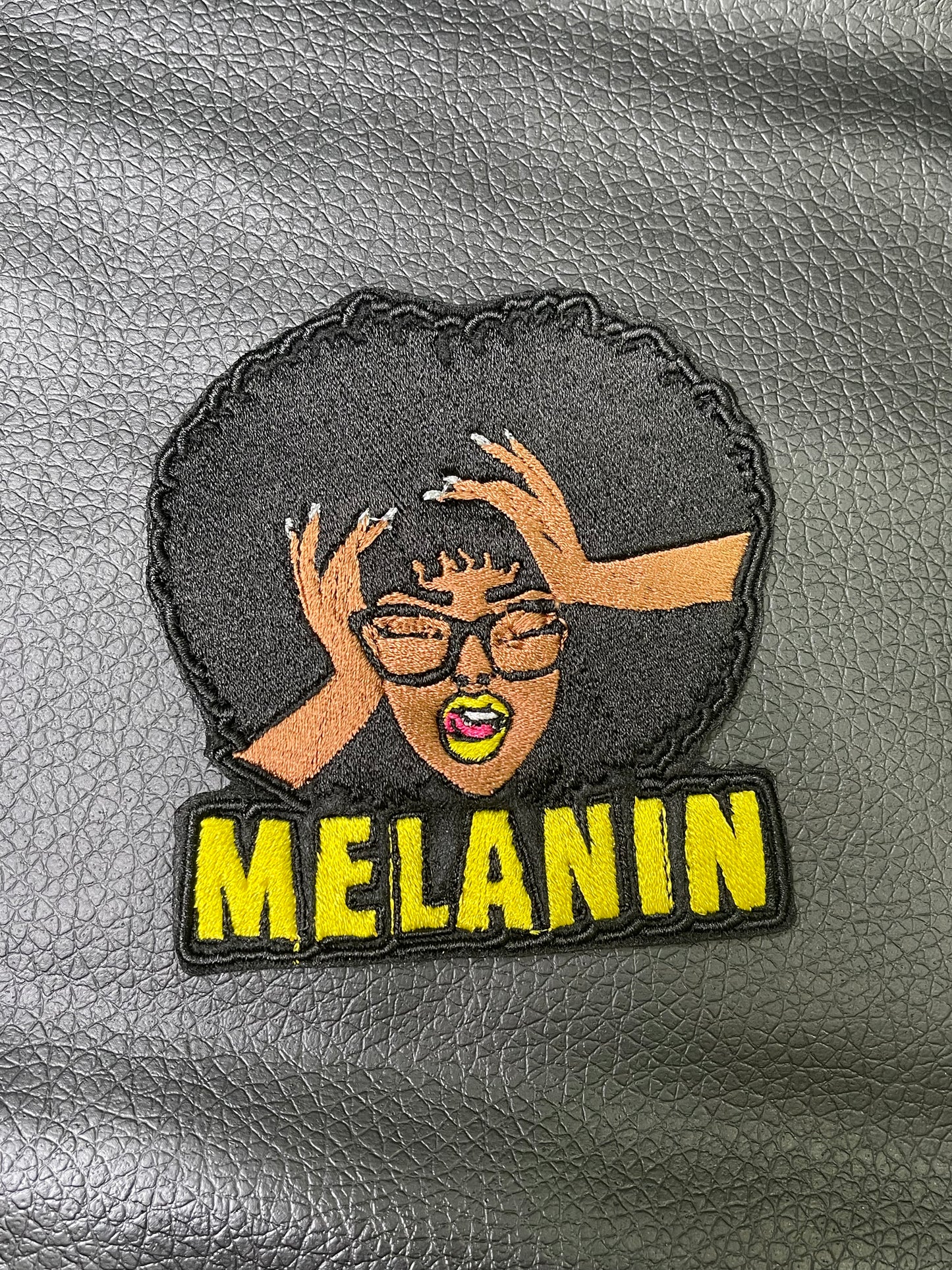 An embroidered patch featuring the word 'MELANIN' and a stylized illustration of a female figure with melonin-rich skin, wearing glasses, and posing with her hands raised.