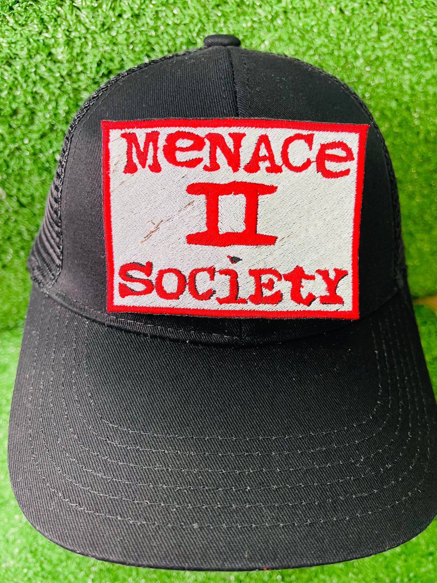 Black cap with 'Menace II Society' patch on a green grass background. Patch with 'Menace II Society' text on green grass. Squared embroidered iron-on patch with red border and red text reading Menace II Society on a white background.