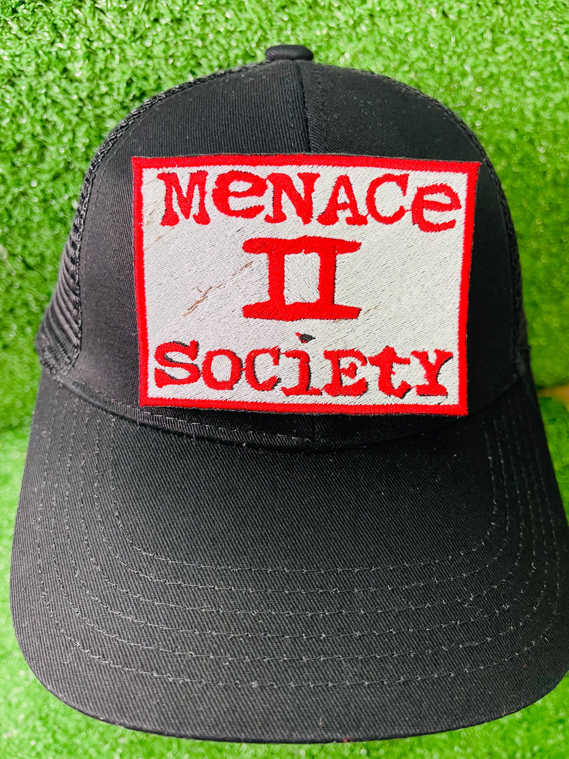 Black cap with 'Menace II Society' patch on a green grass background. Patch with 'Menace II Society' text on green grass. Squared embroidered iron-on patch with red border and red text reading Menace II Society on a white background.