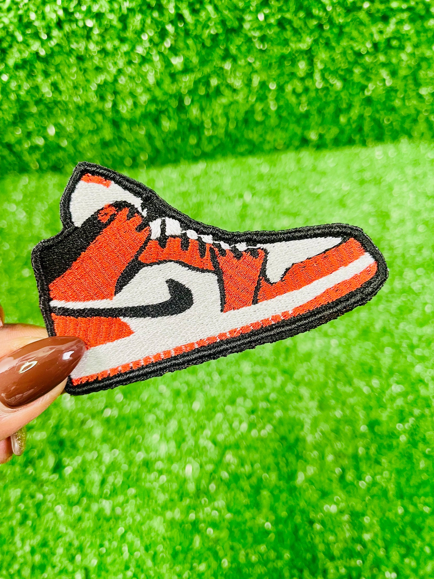 Embroidered patch of a sneaker with orange, black, and white design held over green fabric.