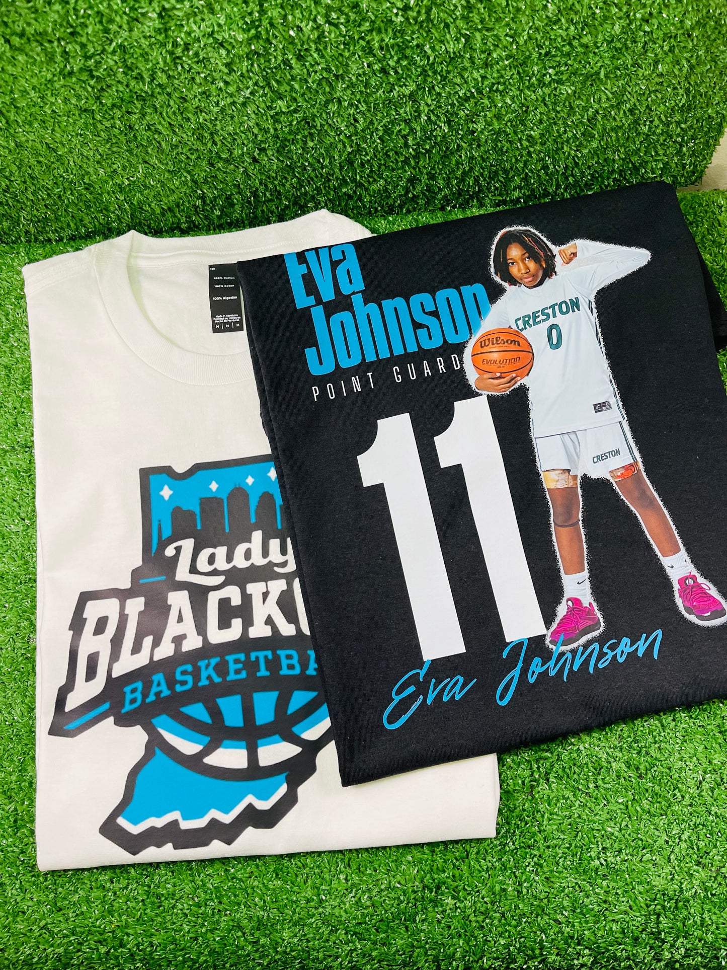 Two custom shirts side by side one white and one black featuring the bold Play Like a Girl graphic on the back displayed on turf with Rare Tagz logo.