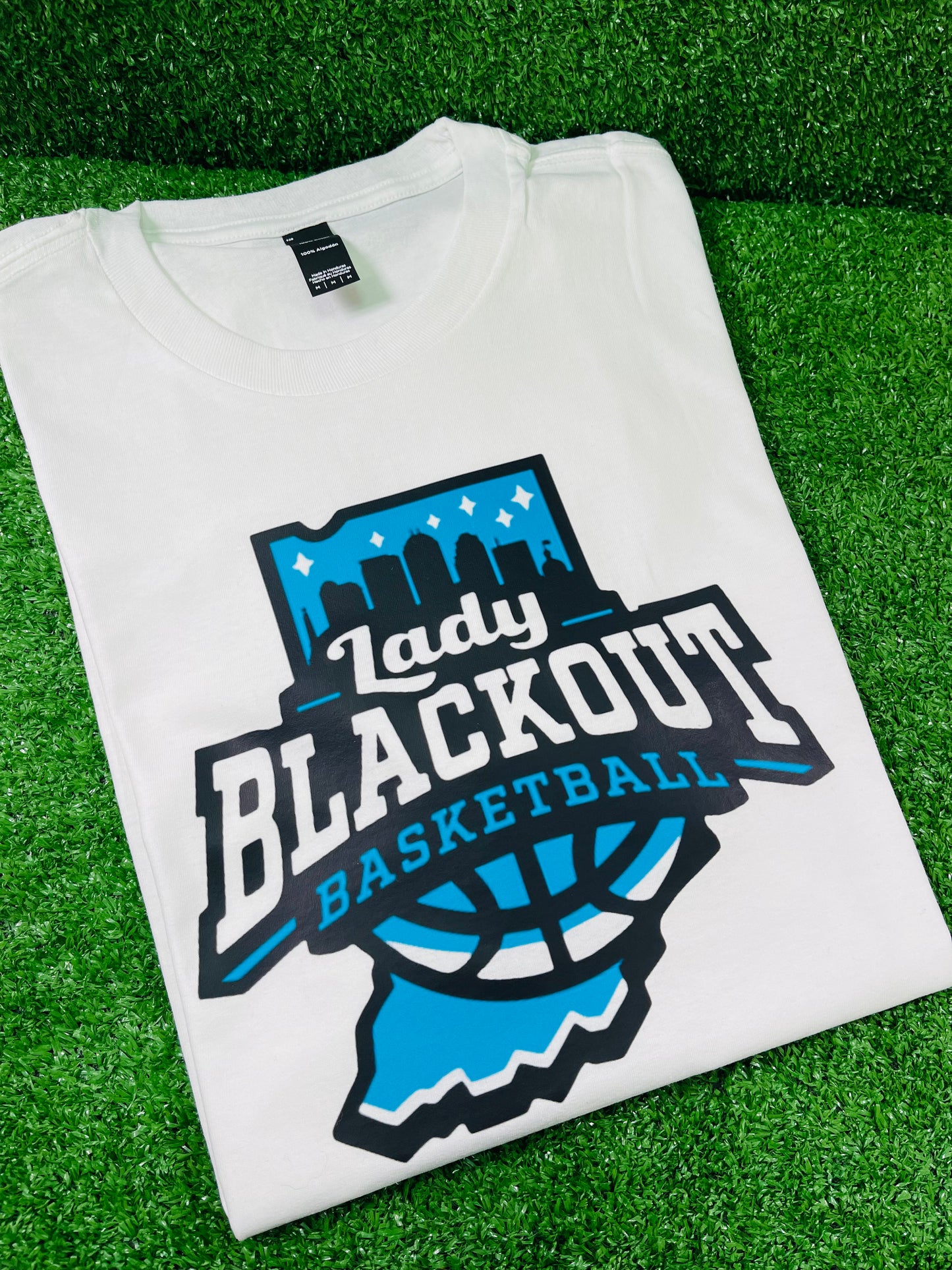 Custom white team shirt featuring Lady Blackout basketball logo in blue and black, displayed on green turf with Rare Tagz branding. Customization is available.