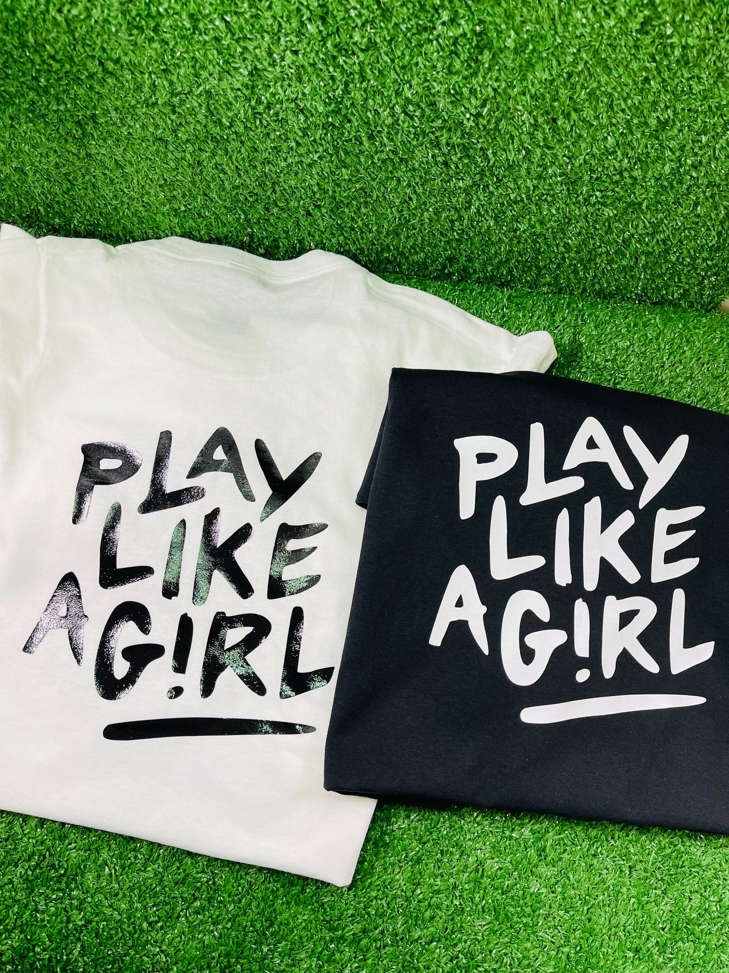 Two custom shirts side by side one white and one black featuring the bold Play Like a Girl graphic on the back displayed on turf with Rare Tagz logo.