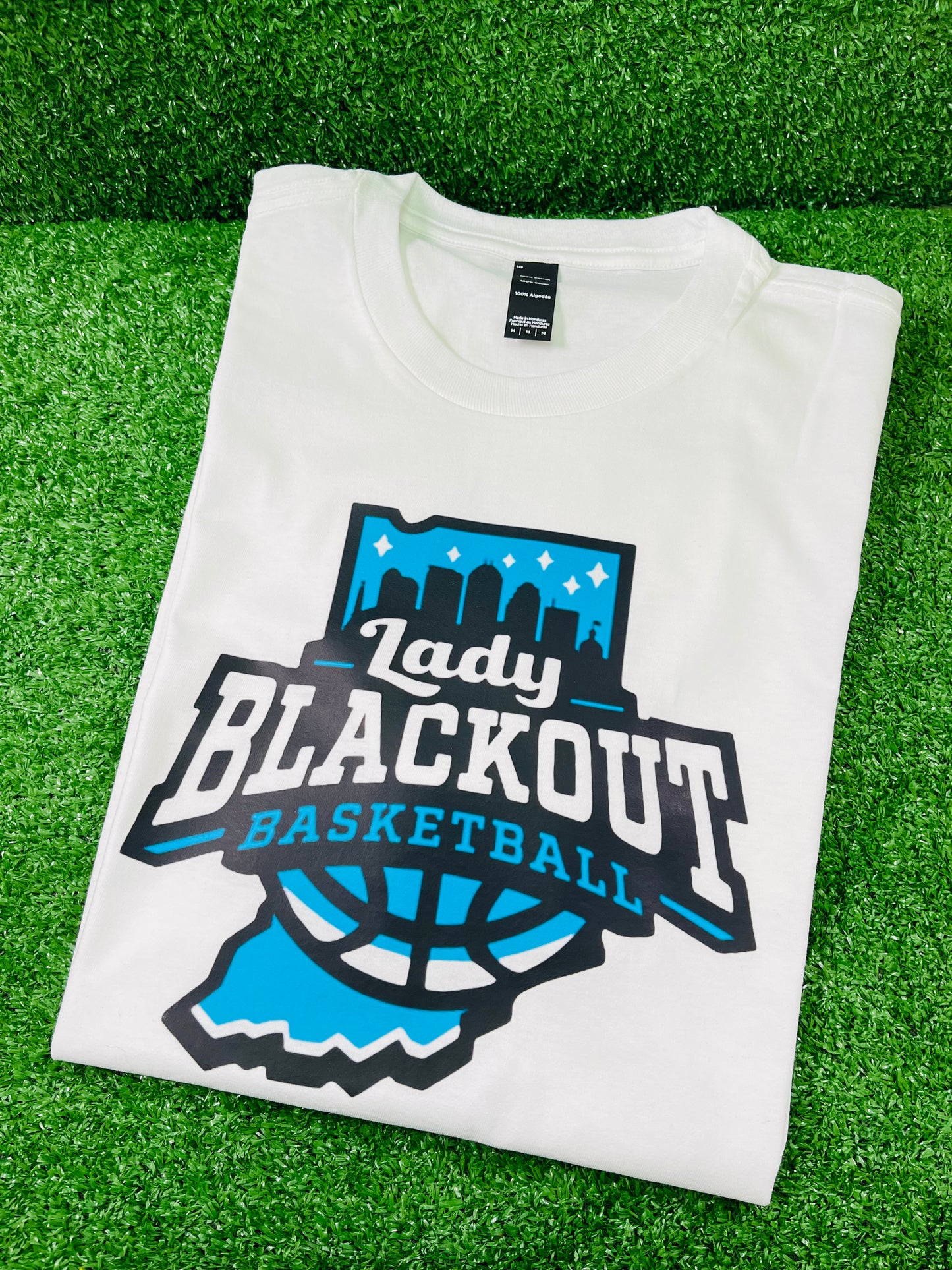 Custom white team shirt featuring Lady Blackout basketball logo in blue and black, displayed on green turf with Rare Tagz branding. Customization is available.