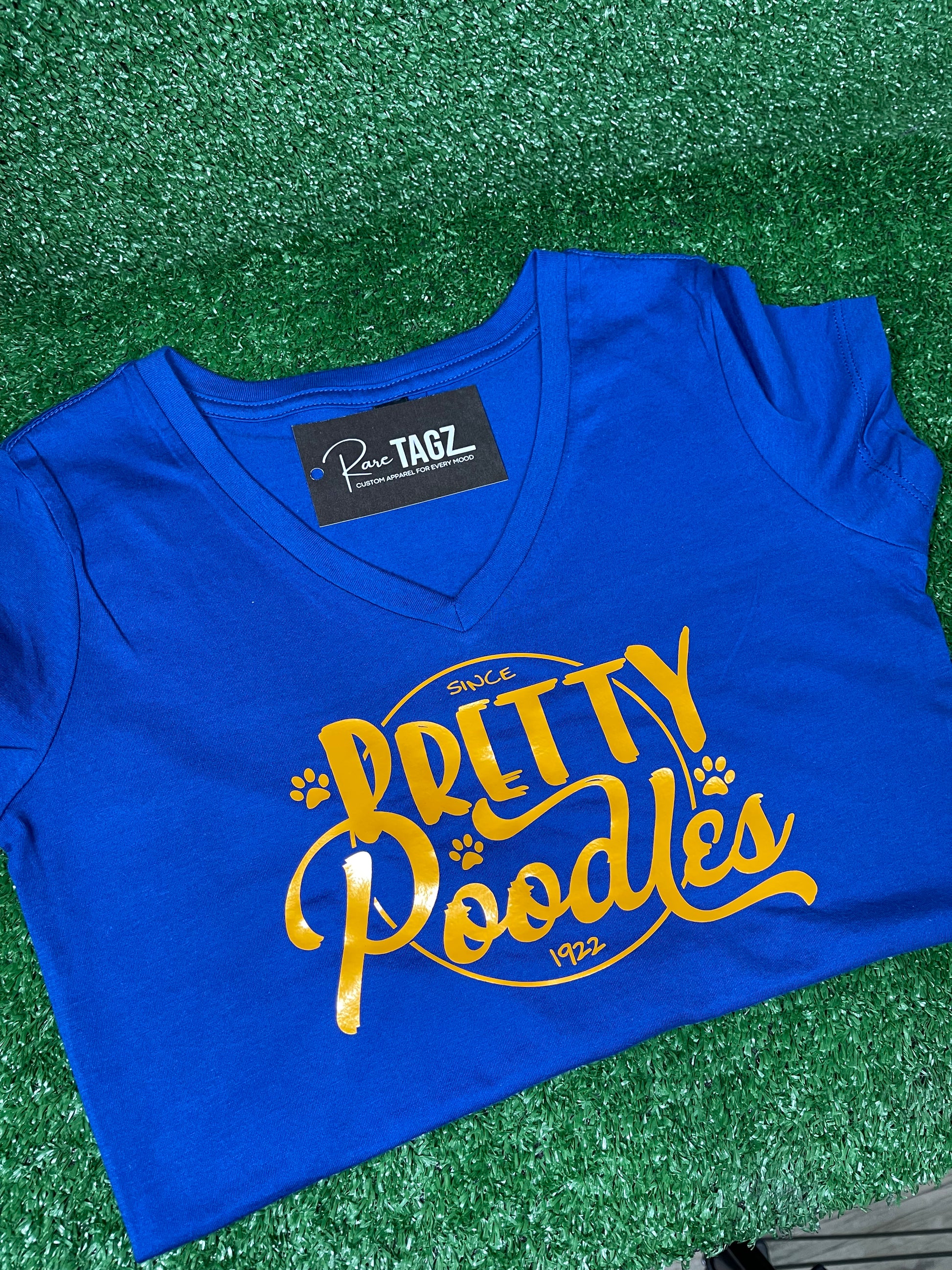 A vibrant blue shirt featuring a stylish 'Pretty Poodle' graphic. The text is elegantly displayed in shimmering gold/yellow font, creating a stunning contrast against the blue background. Sigma Gamma Rho Sorority