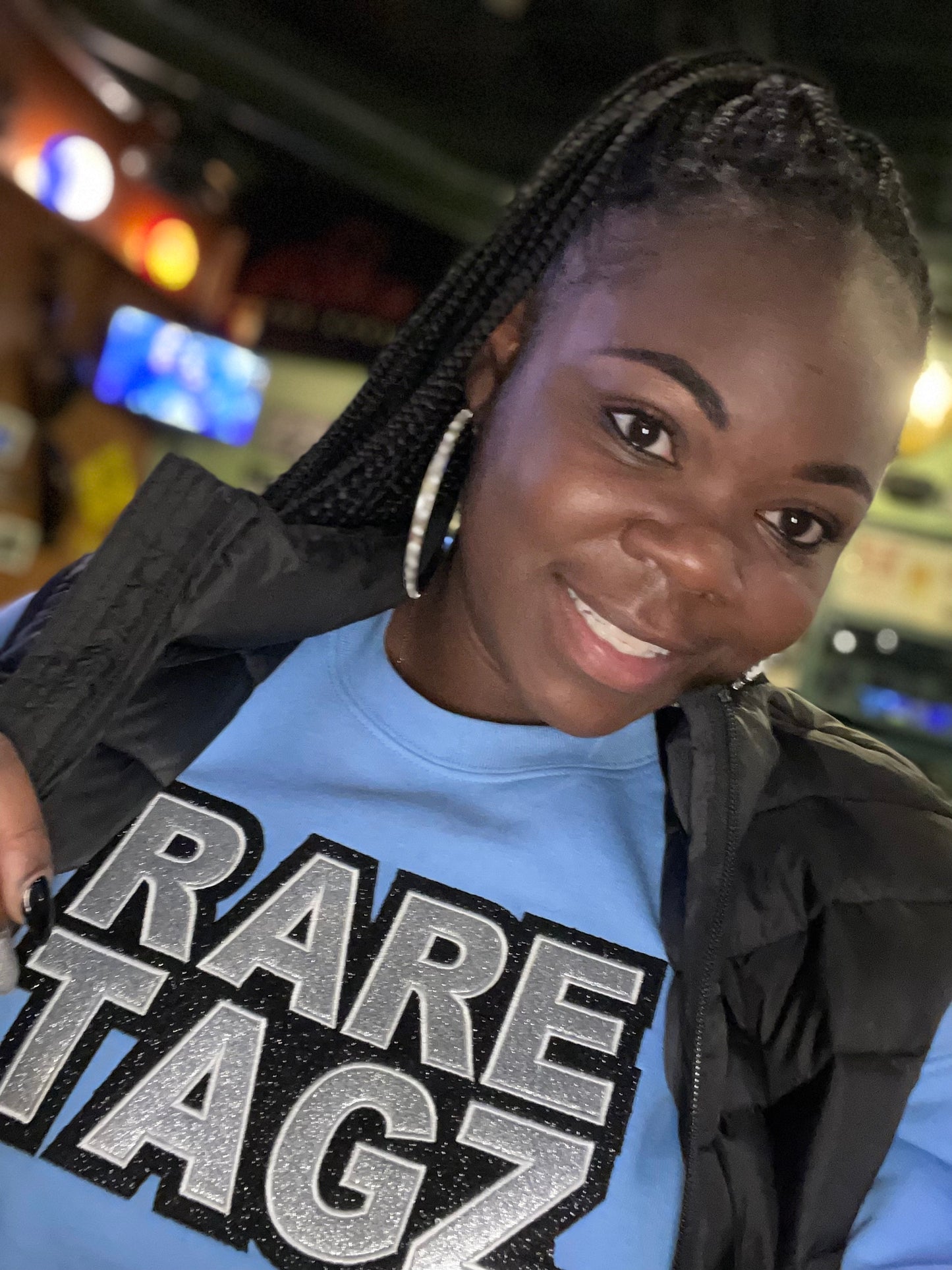 A smiling woman wearing a light blue sweatshirt with the bold, glittery logo Rare Tagz prominently displayed.