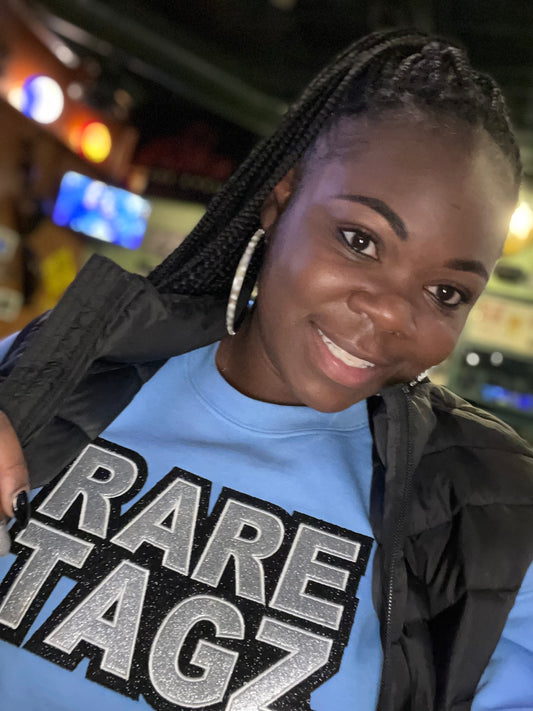 A smiling woman wearing a light blue sweatshirt with the bold, glittery logo Rare Tagz prominently displayed.