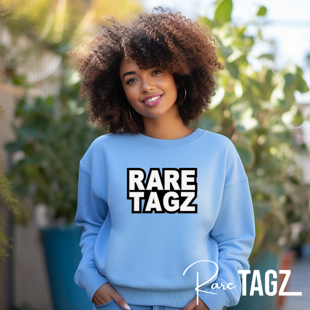 A smiling woman wearing a light blue sweatshirt with the bold, glittery logo Rare Tagz prominently displayed.