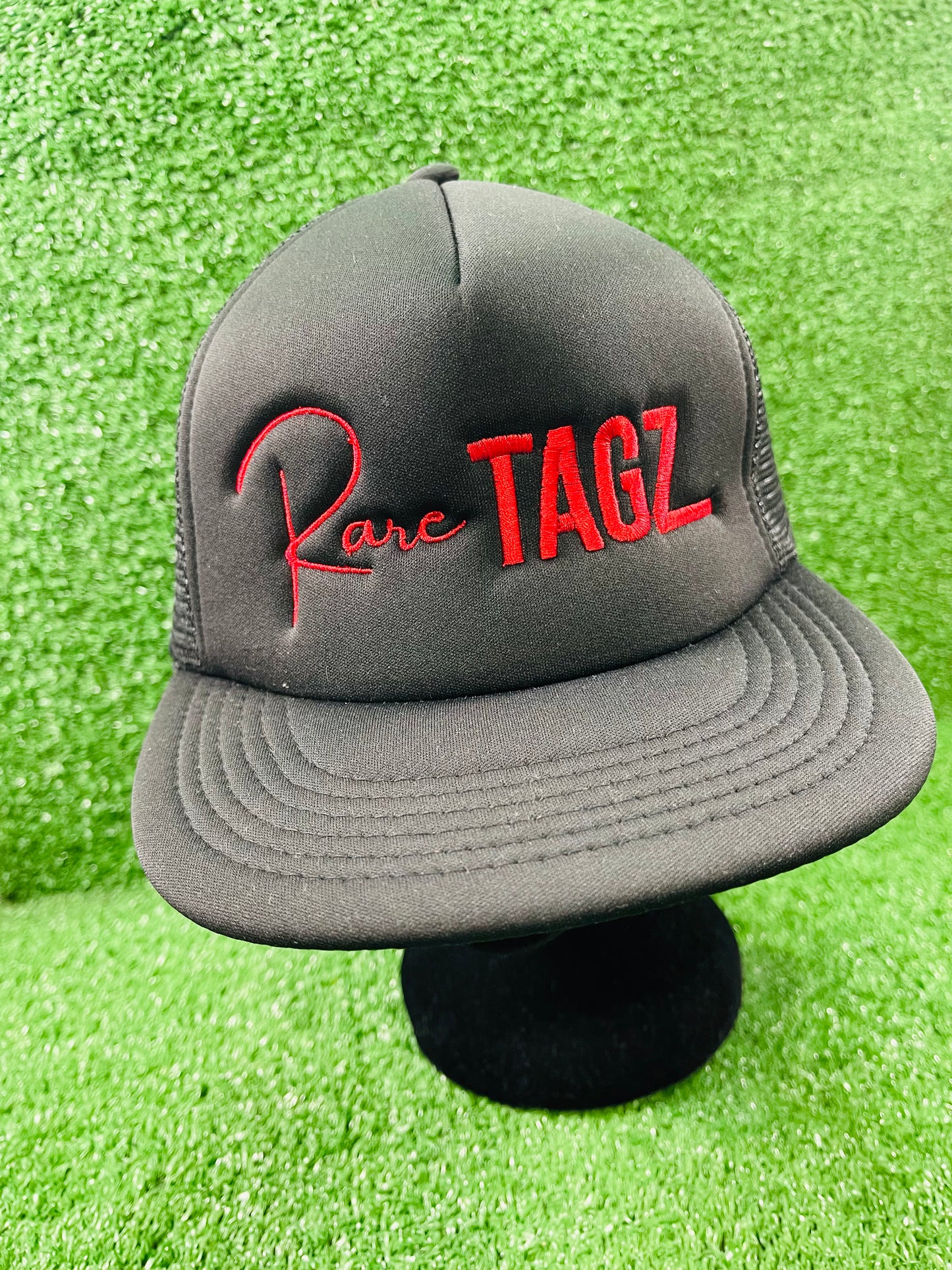 Front view of black trucker hat with red "Rare Tagz" embroidery. Side angle of hat showing clean stitching and mesh back.