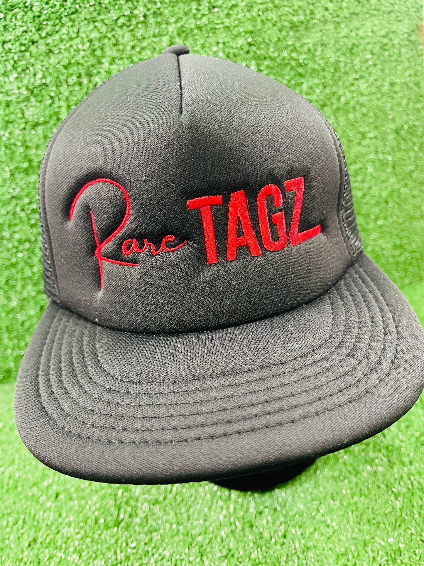 Front view of black trucker hat with red "Rare Tagz" embroidery. Side angle of hat showing clean stitching and mesh back.