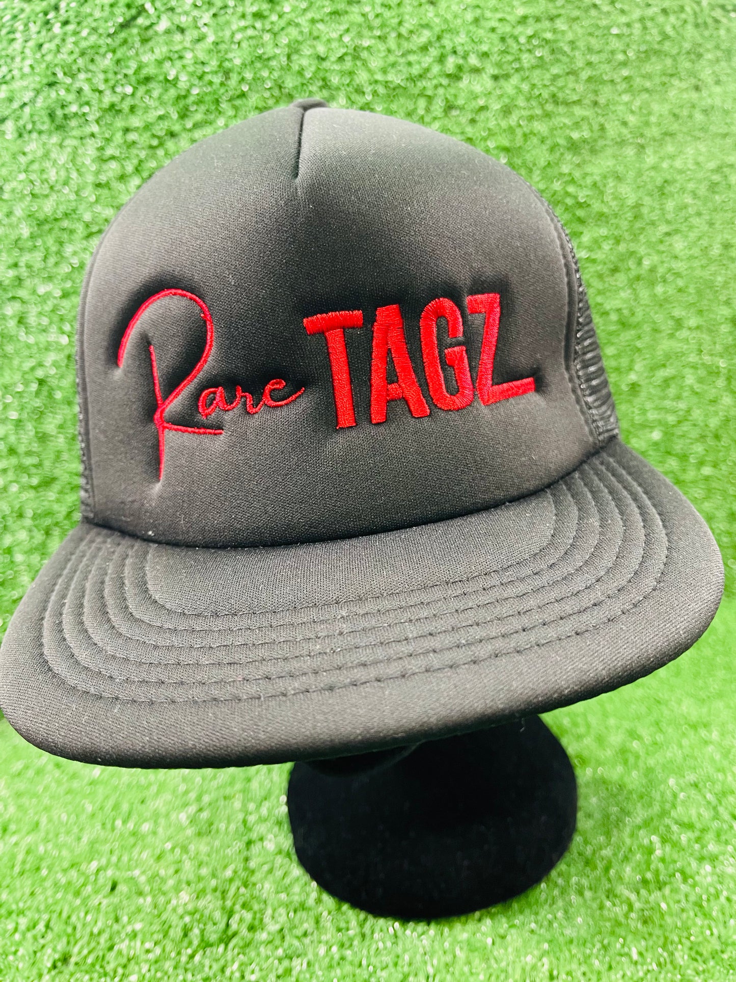 Front view of black trucker hat with red "Rare Tagz" embroidery. Side angle of hat showing clean stitching and mesh back.