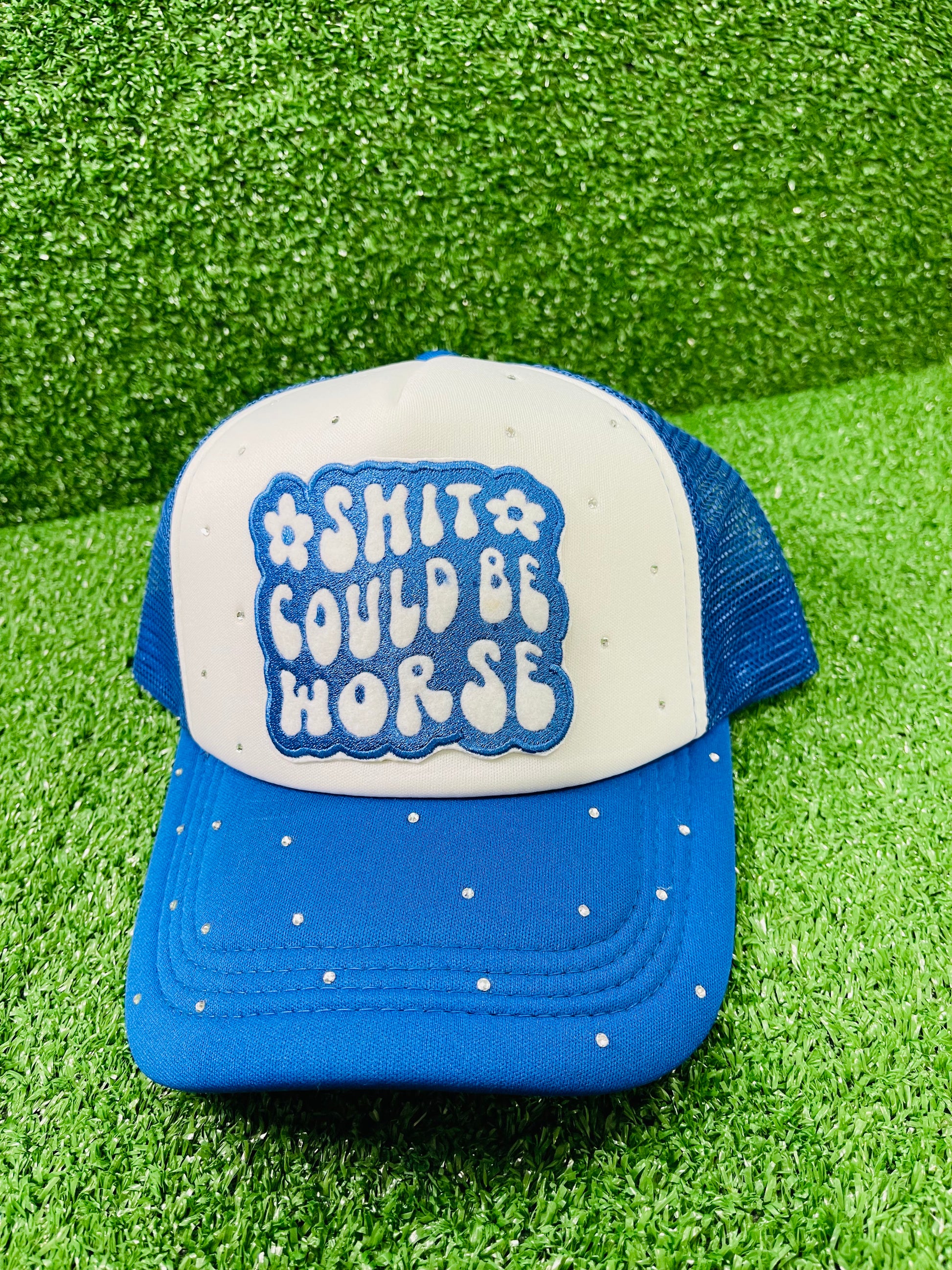 Blue and White custom trucker hat with a bold embroidered patch that reads Shit Could Be Worse and hand-placed rhinestone embellishments on the crown and brim, displayed on a green turf background.