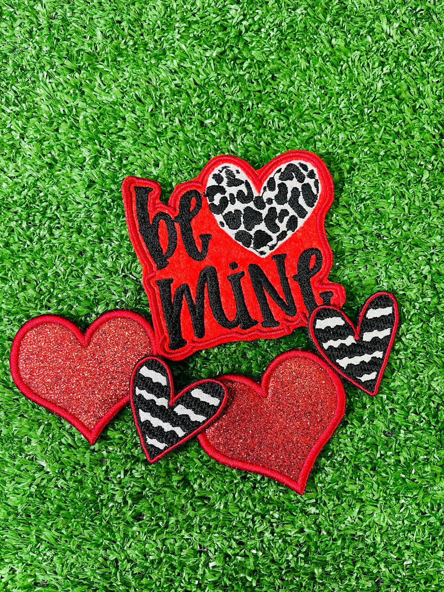 Valentine's Day patch set featuring a bold Be Mine design with a leopard heart, two red glitter hearts, and two black and white zebra stripped hearts displayed on a green grass background.