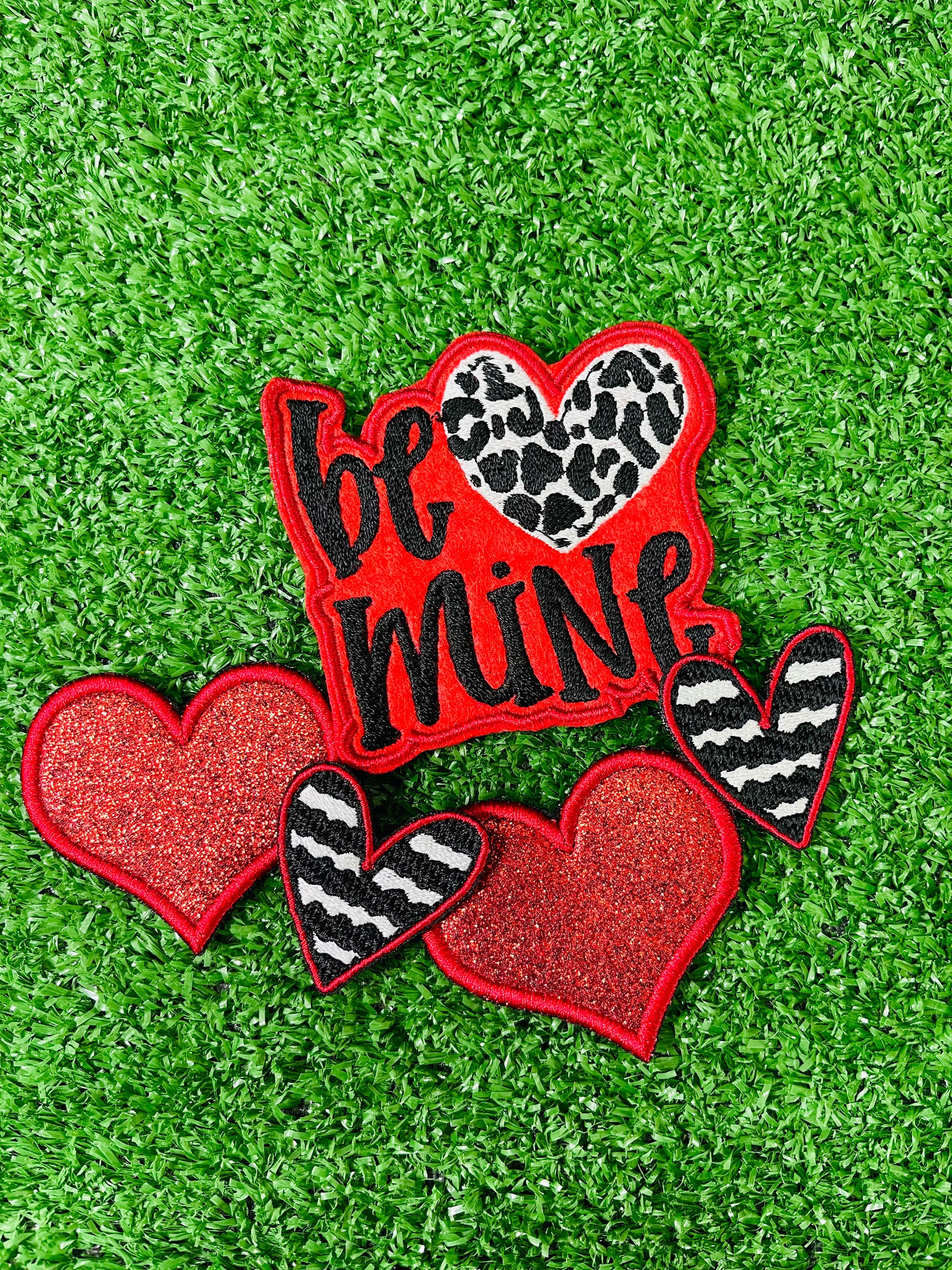 Valentine's Day patch set featuring a bold Be Mine design with a leopard heart, two red glitter hearts, and two black and white zebra stripped hearts displayed on a green grass background.