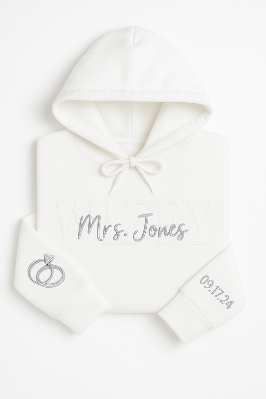 White custom embroidered hoodie with WIFEY and Mrs. Foster stitched on the front, wedding rings embroidered on one sleeve and wedding date on the other, displayed on a white background.