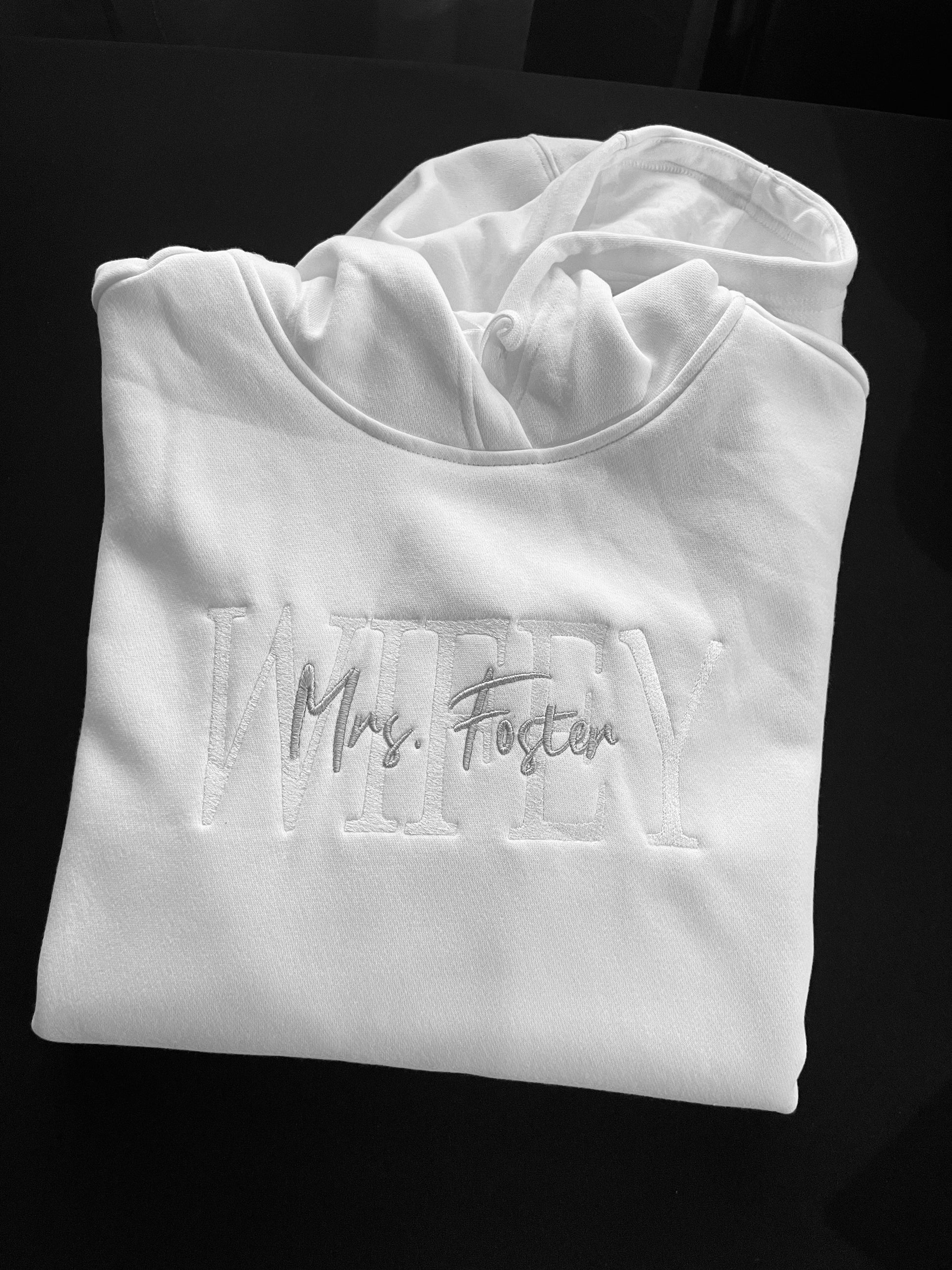 White custom embroidered hoodie with WIFEY and Mrs. Foster stitched on the front, wedding rings embroidered on one sleeve and wedding date on the other, displayed black background.
