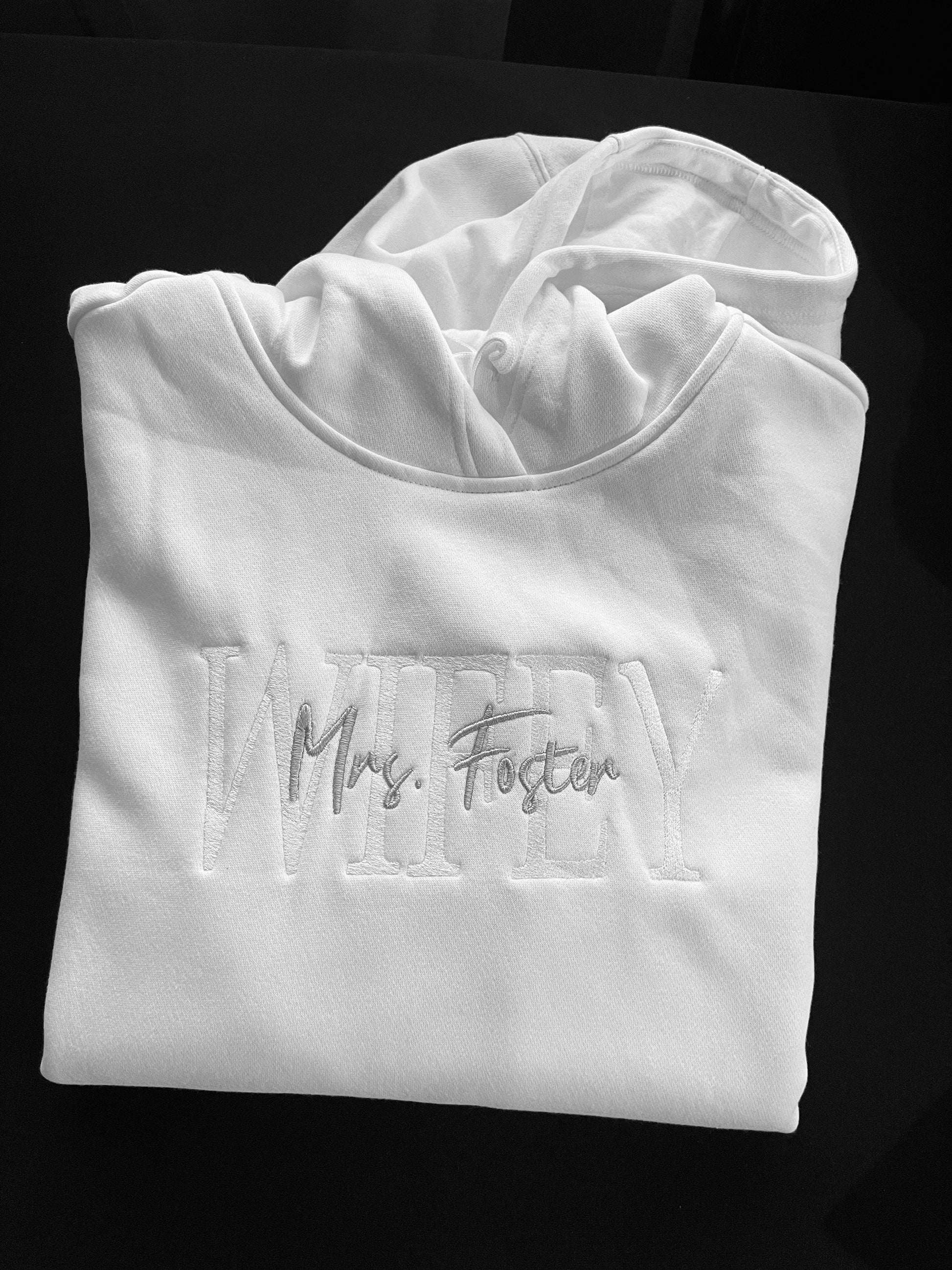 White custom embroidered hoodie with WIFEY and Mrs. Foster stitched on the front, wedding rings embroidered on one sleeve and wedding date on the other, displayed black background.