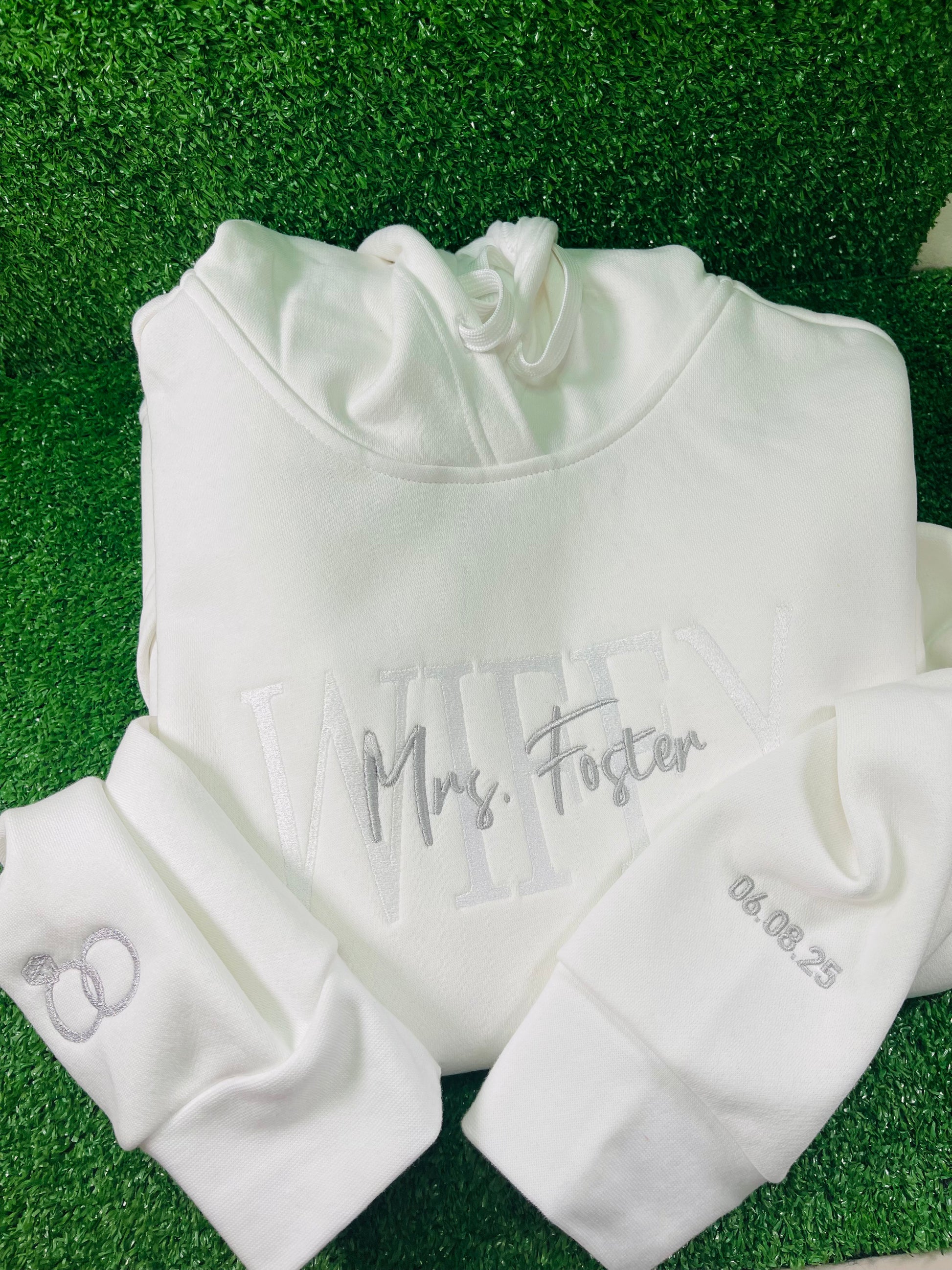 White custom embroidered hoodie with WIFEY and Mrs. Foster stitched on the front, wedding rings embroidered on one sleeve and wedding date on the other, displayed black background.