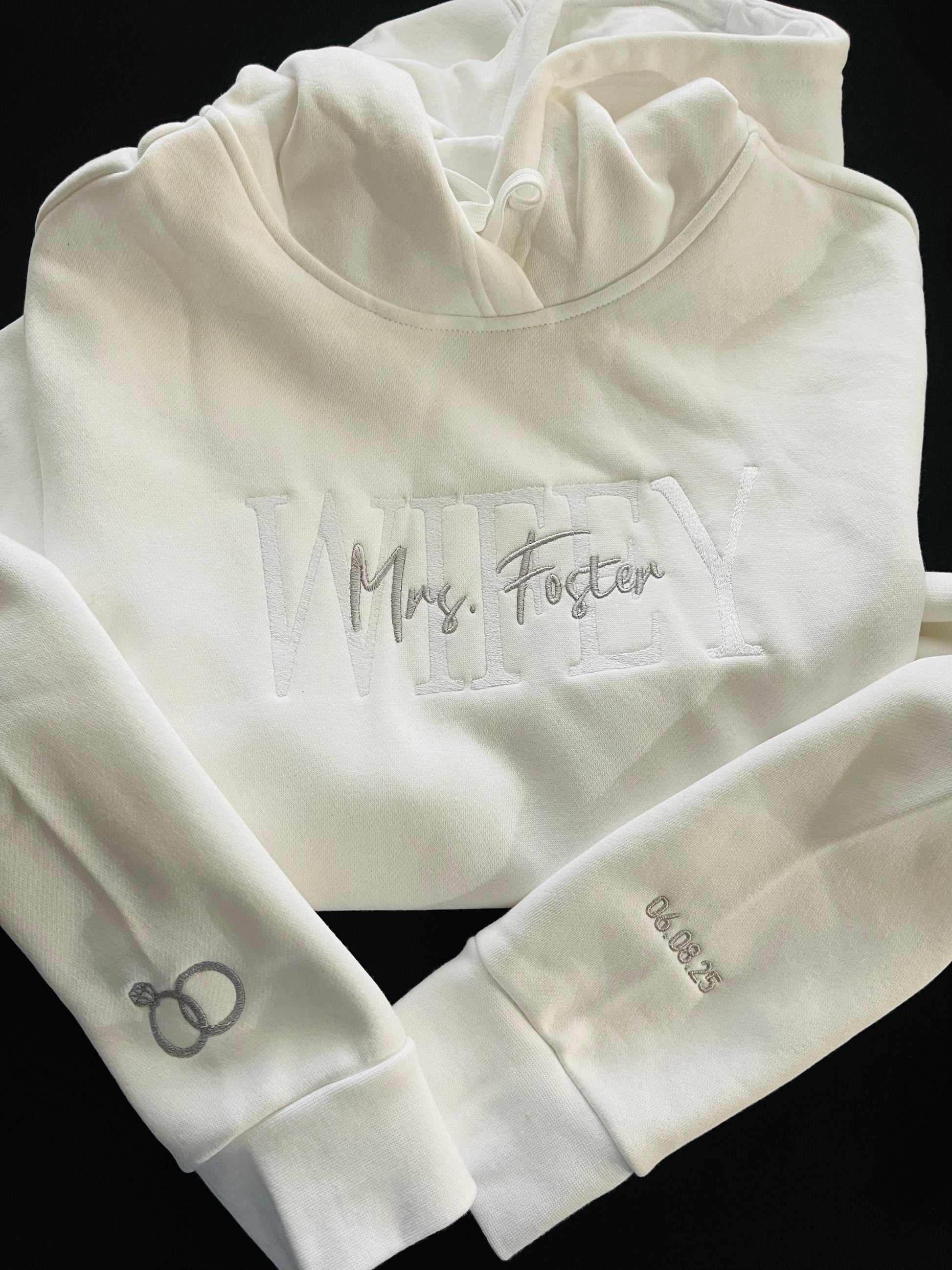 White custom embroidered hoodie with WIFEY and Mrs. Foster stitched on the front, wedding rings embroidered on one sleeve and wedding date on the other, displayed black background.